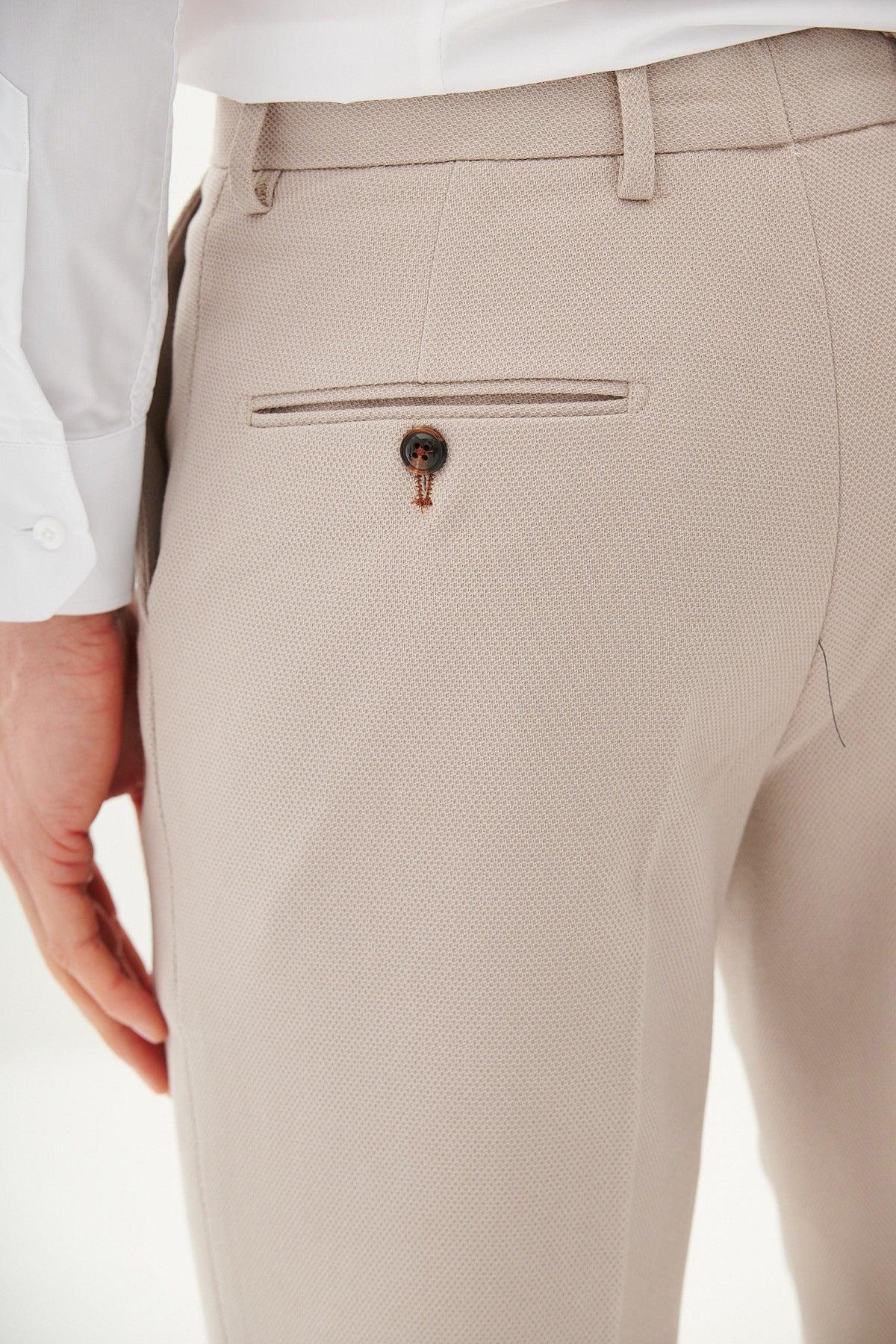 A close-up shows a person wearing marcdarcy's HM5 - Stone Tailored Trousers in light beige, styled with a white shirt. The slim fit pants feature a single buttoned back pocket, and the person's left hand rests relaxed by their side.