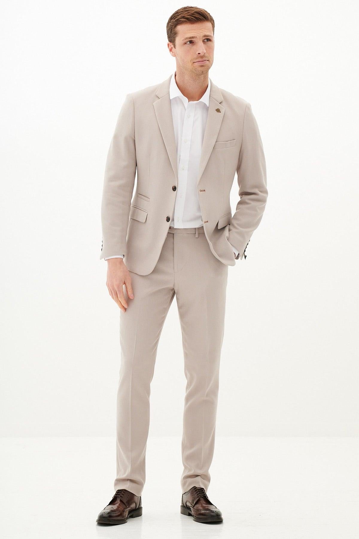 A man stands against a white background, dressed in the Marc Darcy HM5 - Stone Tailored Two Piece Suit with a white shirt and brown dress shoes. His left hand is in his pocket as he gazes slightly to the side with a neutral expression.