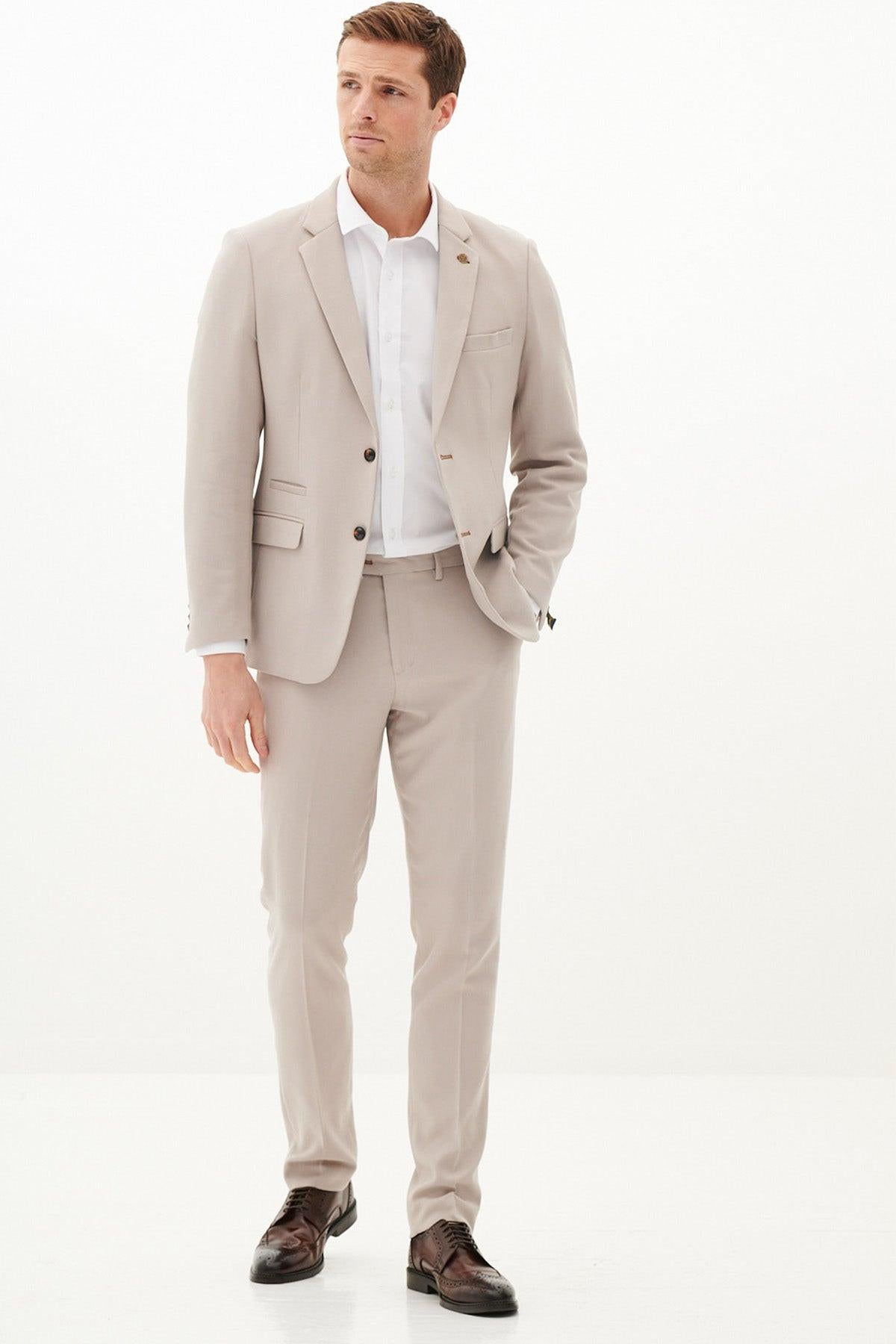 A man wears the Marc Darcy HM5 - Stone Tailored Two Piece Suit, featuring a slim fit in poly-viscose, with a white shirt and brown shoes. He stands against a plain white background, left hand in pocket, looking slightly left.