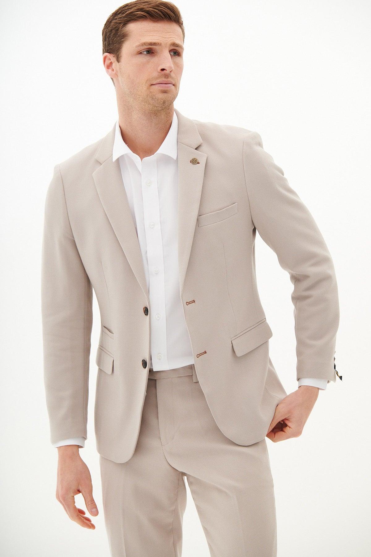 A man models the marcdarcy HM5 - Stone Tailored Blazer in light beige over a white dress shirt, standing against a plain white background and gazing slightly to the side with one hand near his hip.