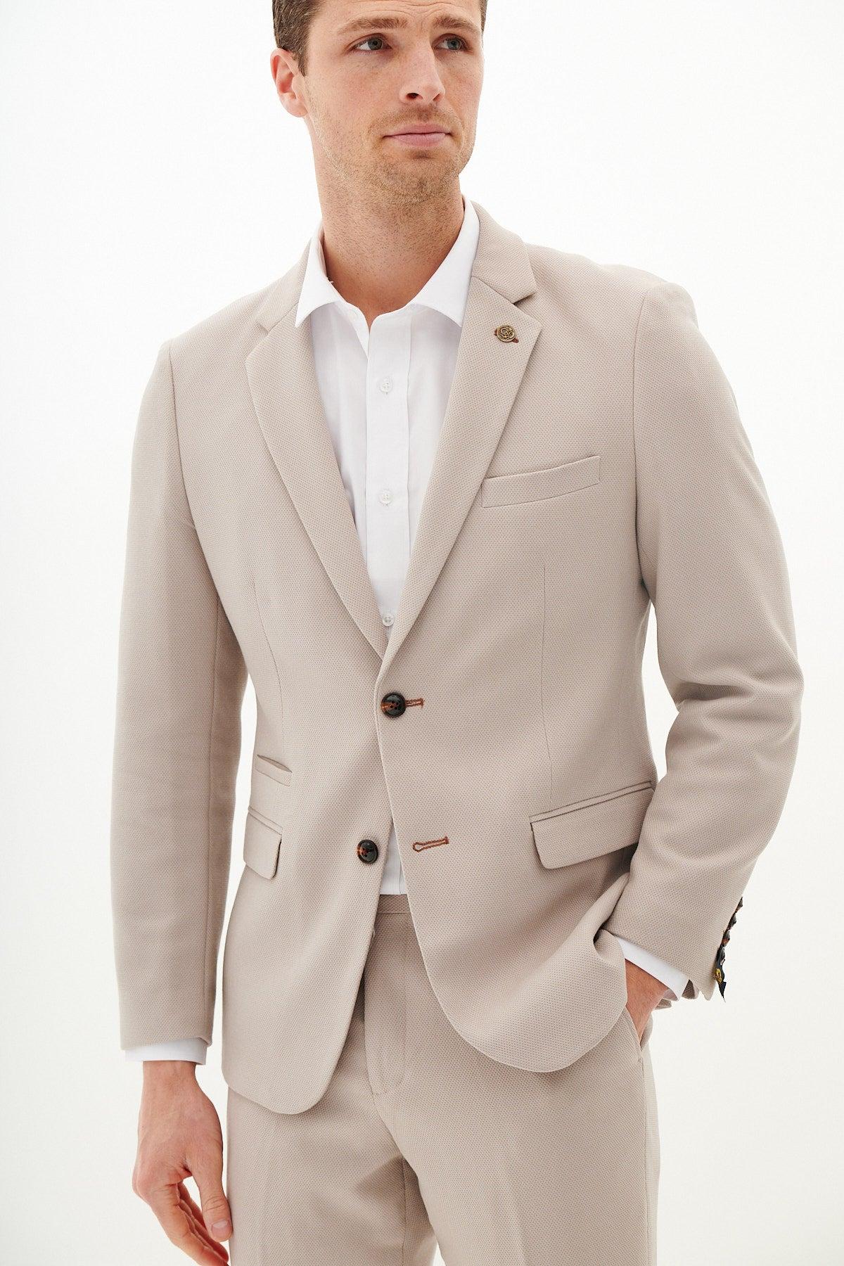 A man wears the Marc Darcy HM5 Stone Blazer with matching tan waistcoat and trousers, featuring notch lapels, a subtle lapel pin, and white dress shirt. Crafted from a comfy lycra blend, he poses with one hand in his pocket against a white background.