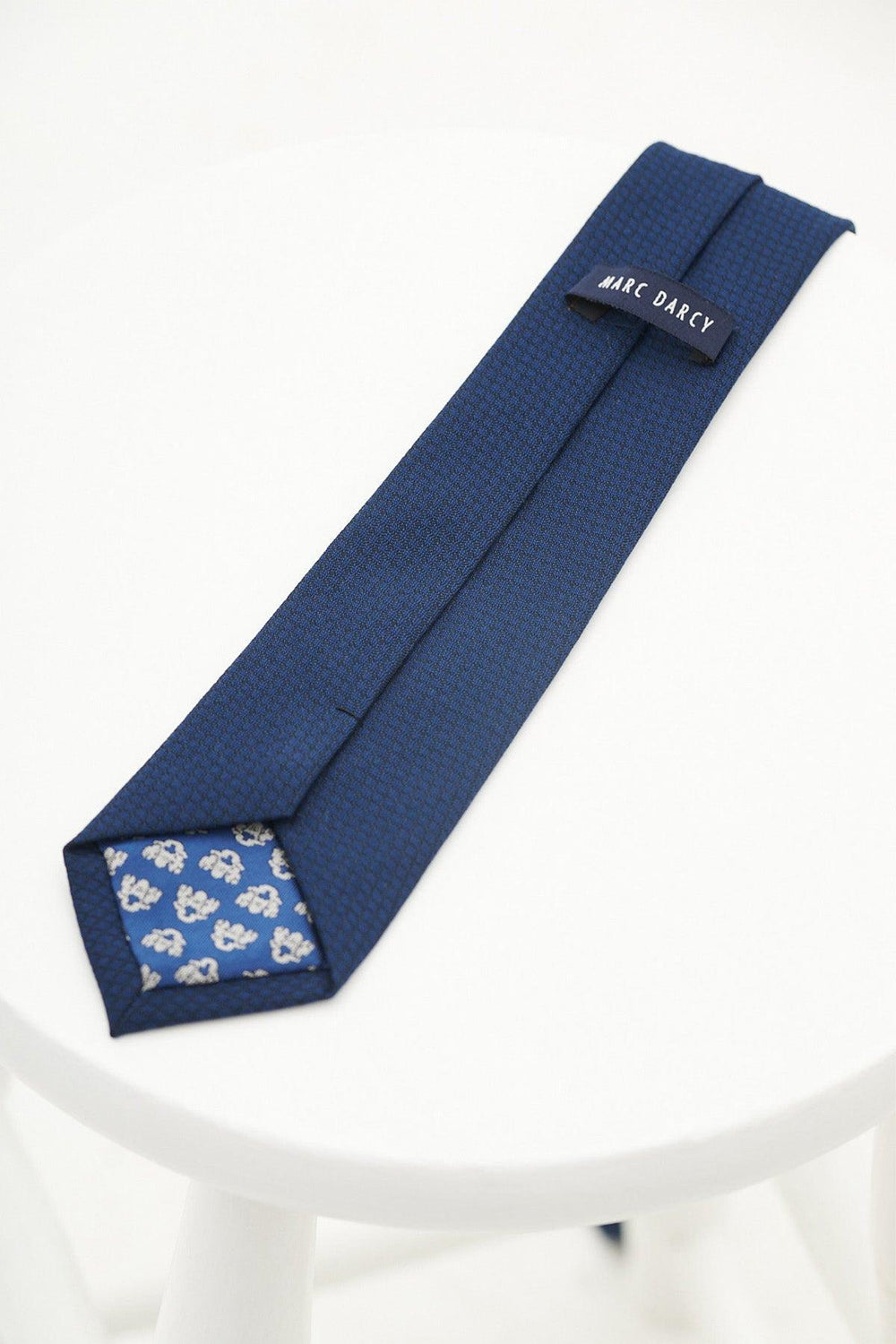 The MAX - Royal Diamond Print Tie by Accessory features a navy diamond pattern, textured finish, and floral inner tip with branding. Displayed on a white round table—ideal for formalwear or weddings.