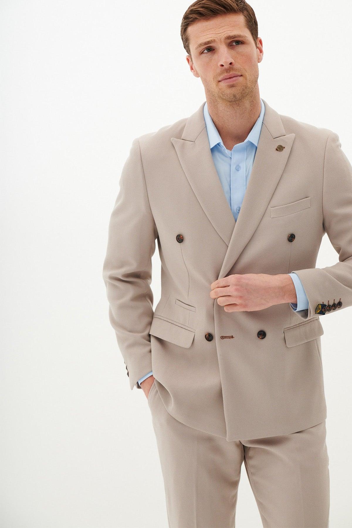 A man wearing the marcdarcy HM5 - Double Breasted Stone Blazer over a light blue dress shirt stands against a plain white background, gazing slightly upward with a neutral expression and adjusting his jacket.