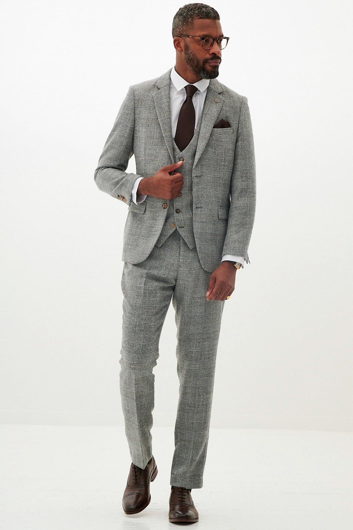 A man in the marcdarcy KRIS - Silver Grey Tweed Suit, paired with a white shirt, dark brown tie, brown shoes, and glasses, stands against a plain white background looking to the side with one hand in his pocket.