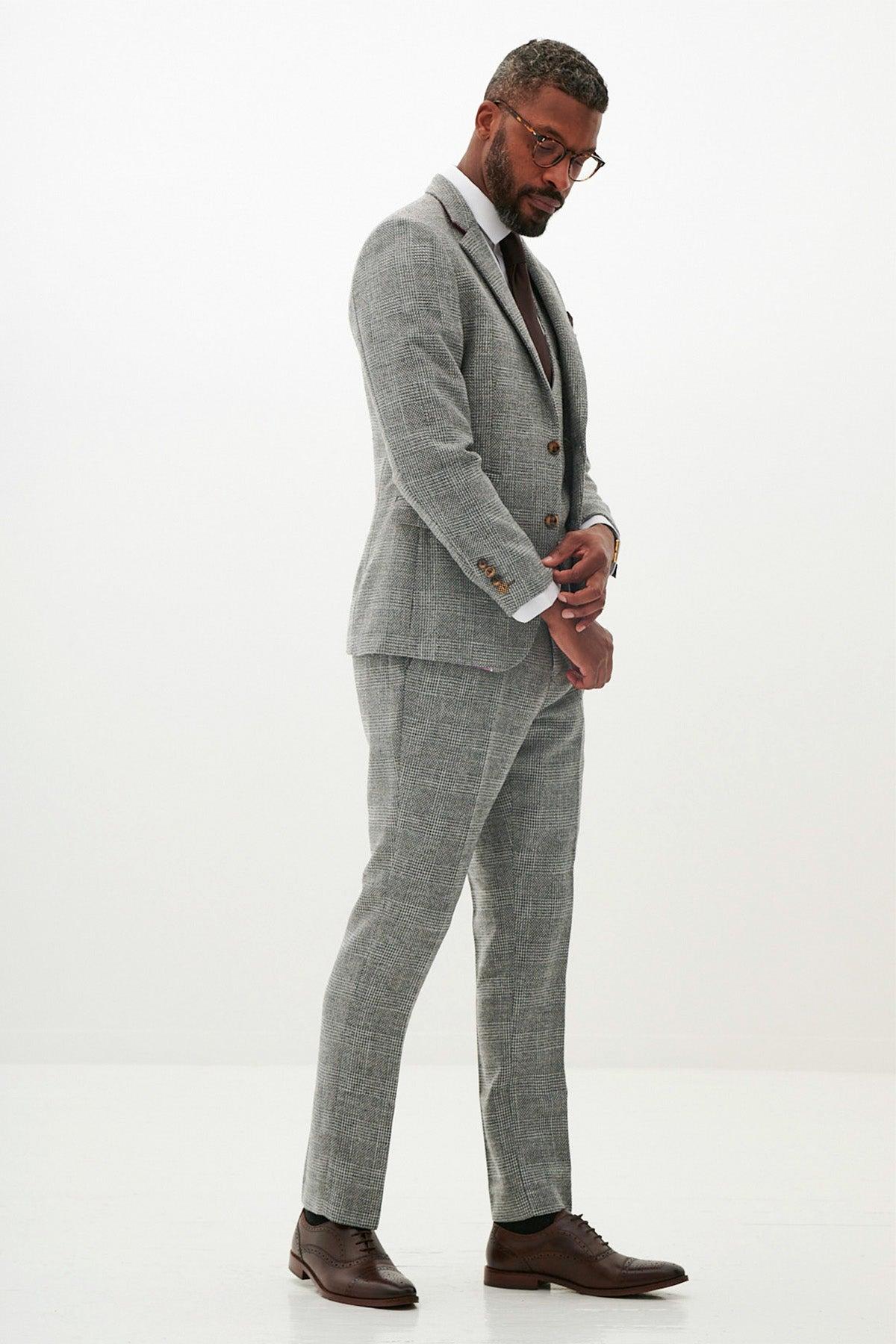 A man in marcdarcy’s WHU Collection - KRIS Silver Grey Tweed Suit, worn by Jarrod Bowen, stands against a white background. He wears a white shirt, brown tie, and brown dress shoes, adjusting his cuff and looking slightly downward.
