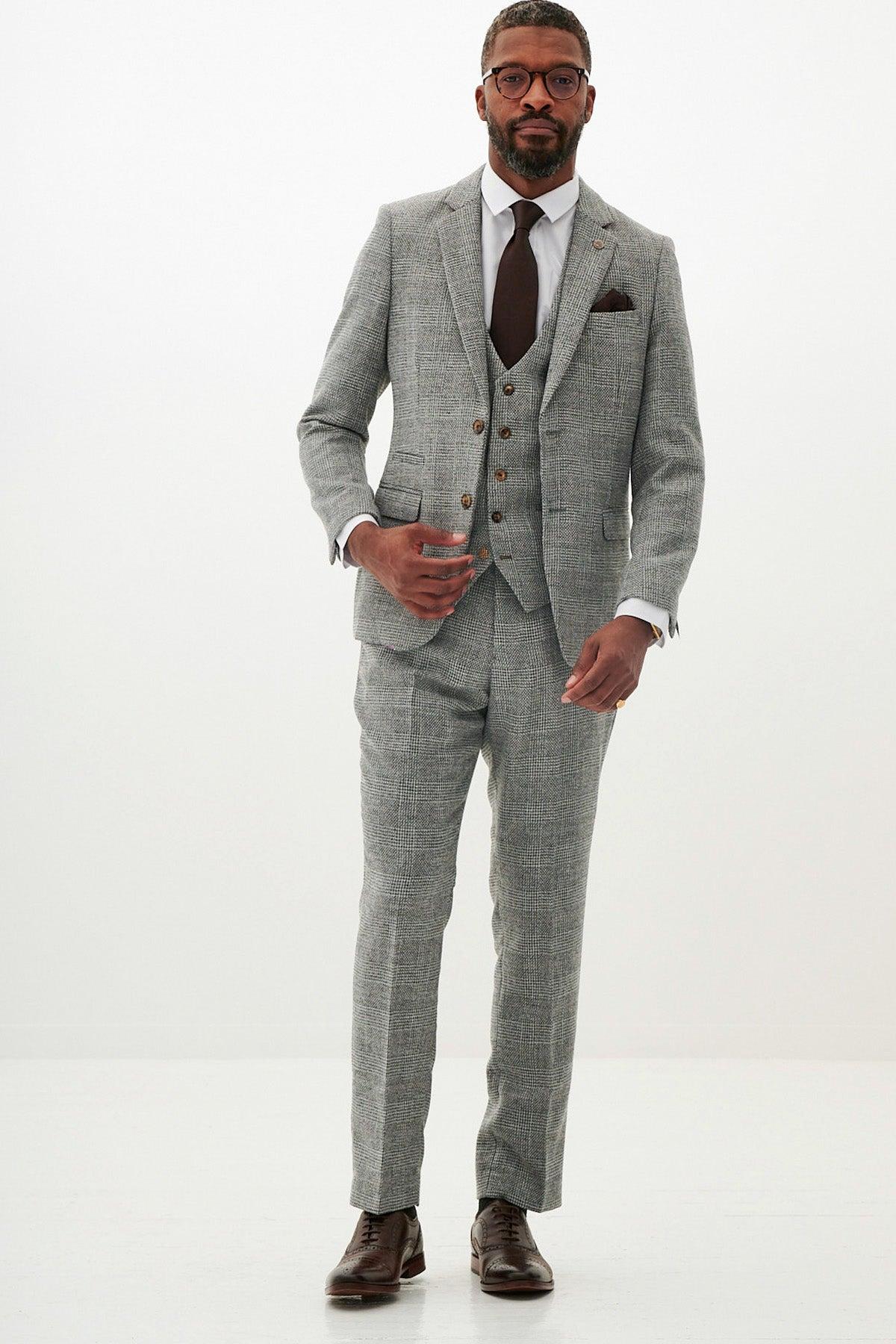 A man wears the marcdarcy KRIS - Silver Grey Tweed Suit, paired with a white shirt, dark brown tie, and brown dress shoes. He has short hair, a trimmed beard, glasses, and stands against a plain white background.
