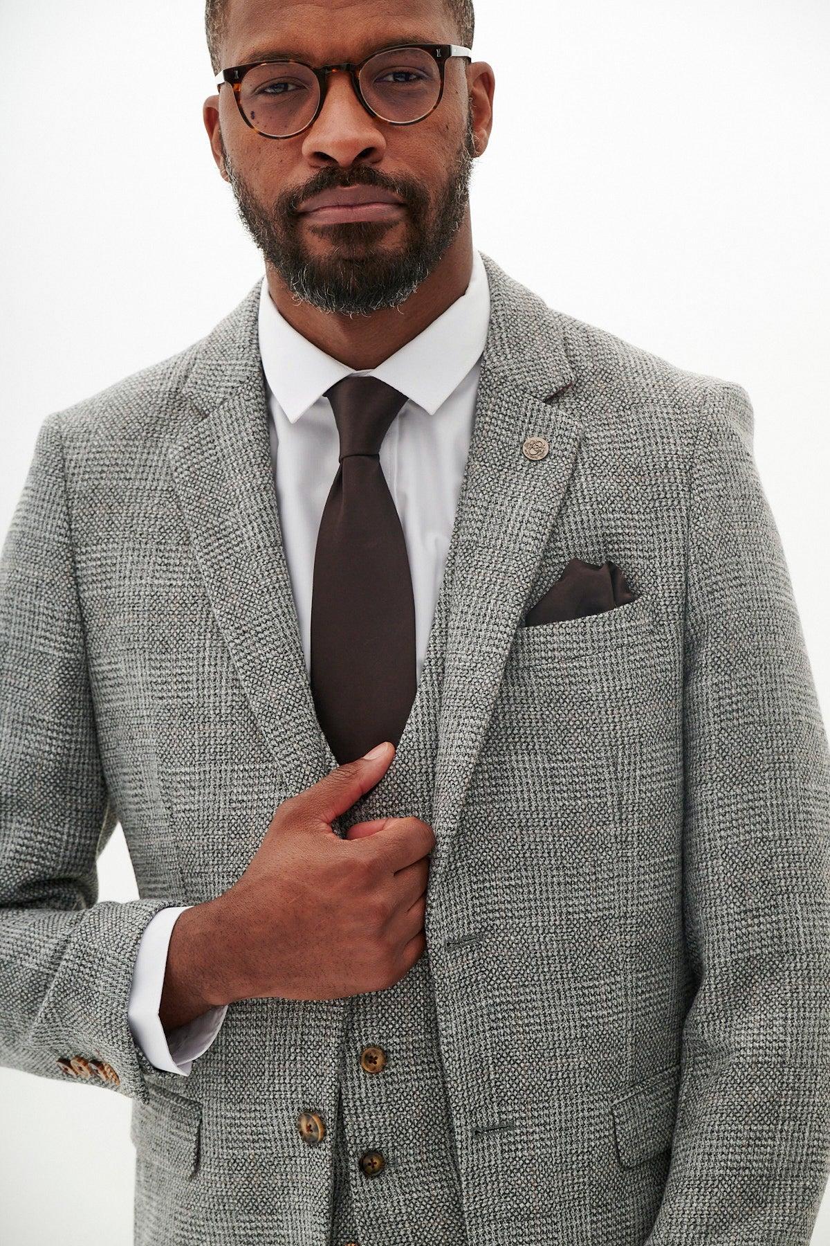 A man in glasses wears the marcdarcy KRIS Silver Grey Tweed Suit with a white shirt, brown tie, and pocket square, standing against a plain white background while holding one side of his jacket.