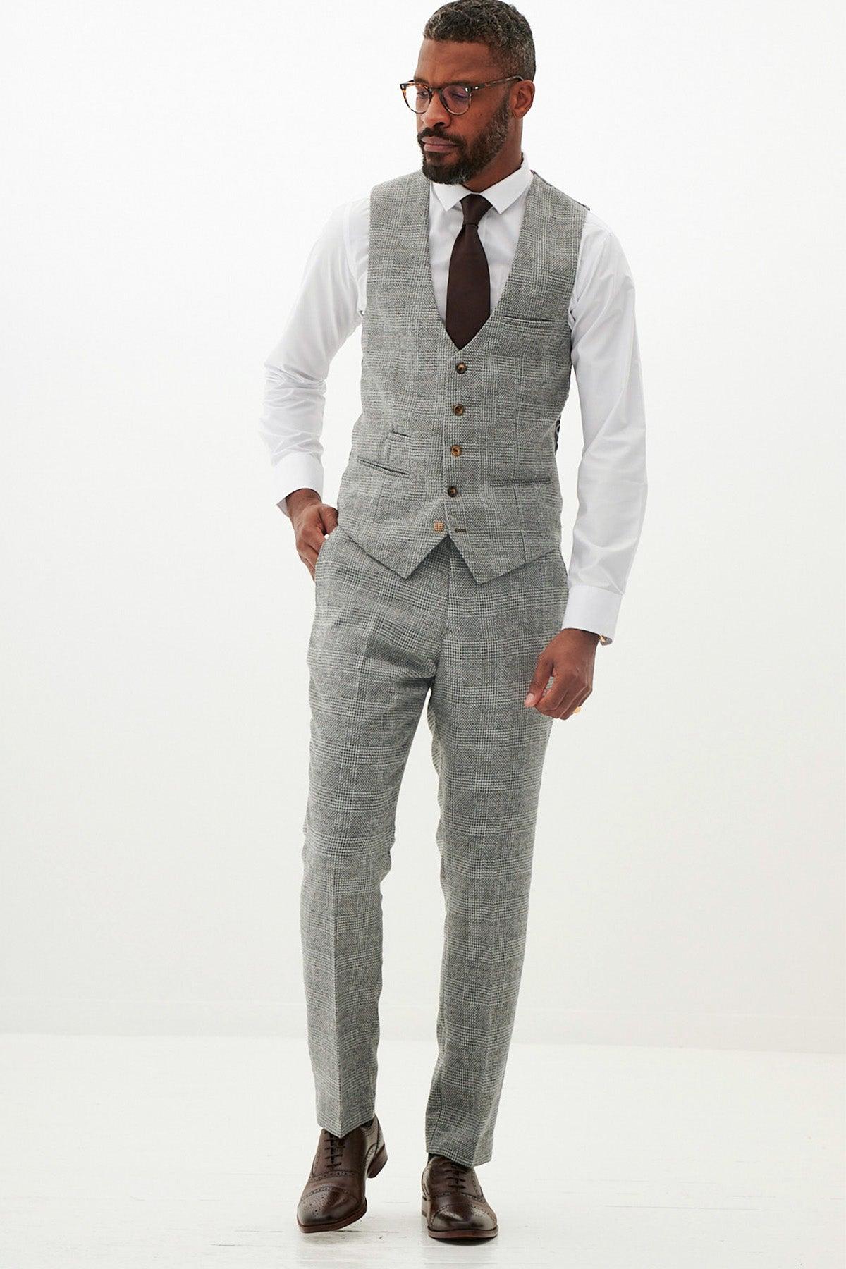 A man wearing glasses, a Marc Darcy KRIS Silver Grey Tweed Waistcoat, matching Prince of Wales check trousers, a white shirt, dark tie, and brown dress shoes stands against a plain white background.