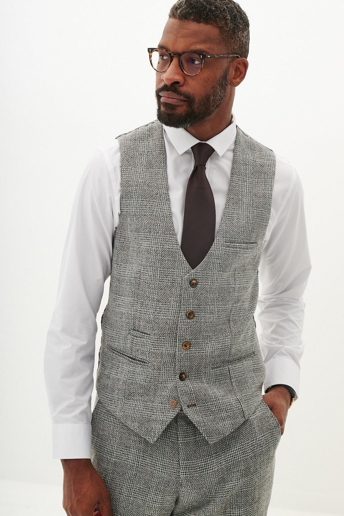 A Black man wearing the Marc Darcy KRIS Silver Grey Tweed Waistcoat, paired with a white shirt, dark tie, and glasses, poses confidently with his hand in his pocket.