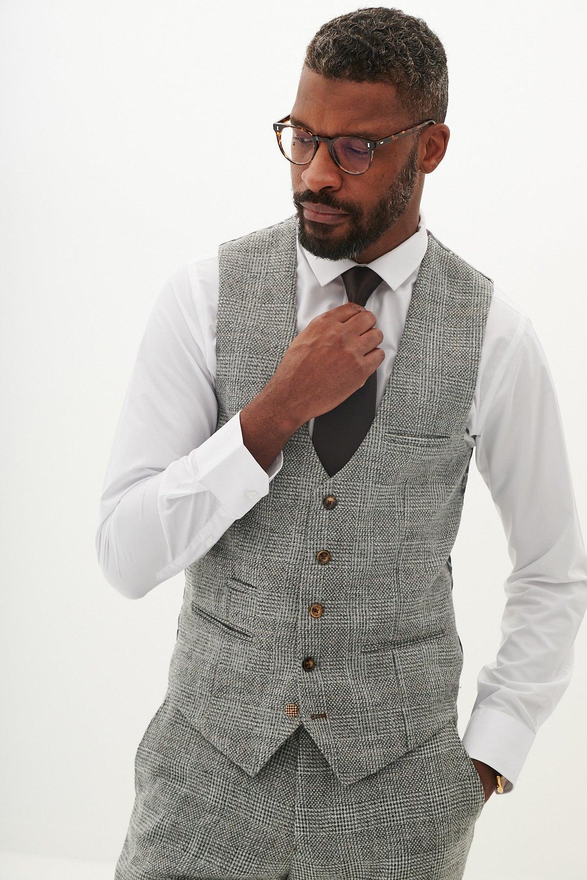 A man wearing glasses, a white dress shirt, dark tie, and the Marc Darcy KRIS - Silver Grey Tweed Waistcoat with matching pants stands against a plain white background, adjusting his tie and looking down.