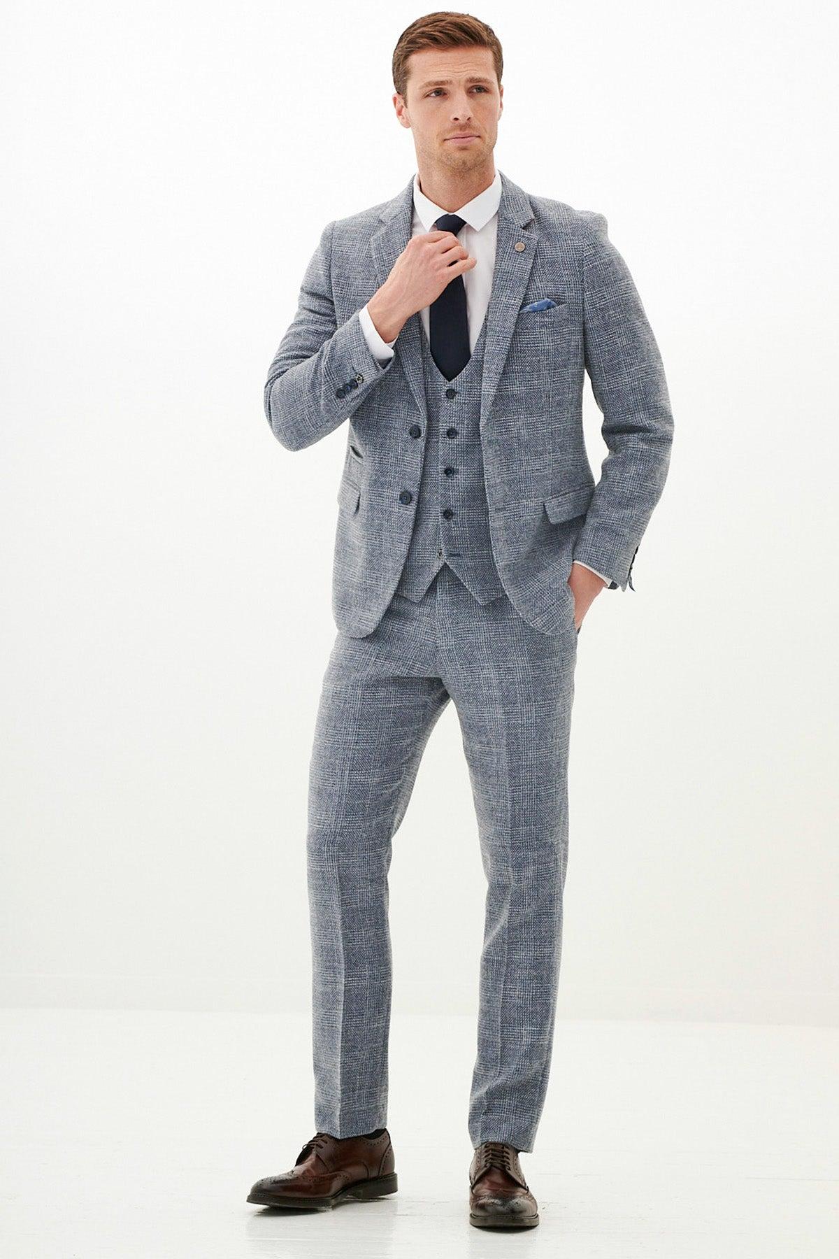 A man in a marcdarcy Official West Bromwich Albion F.C. Club Suit—KRIS Tweed Suit with Kelvin Navy Waistcoat—wears a white shirt, navy tie, blue pocket square, adjusts his tie, and stands with one hand in his pocket.