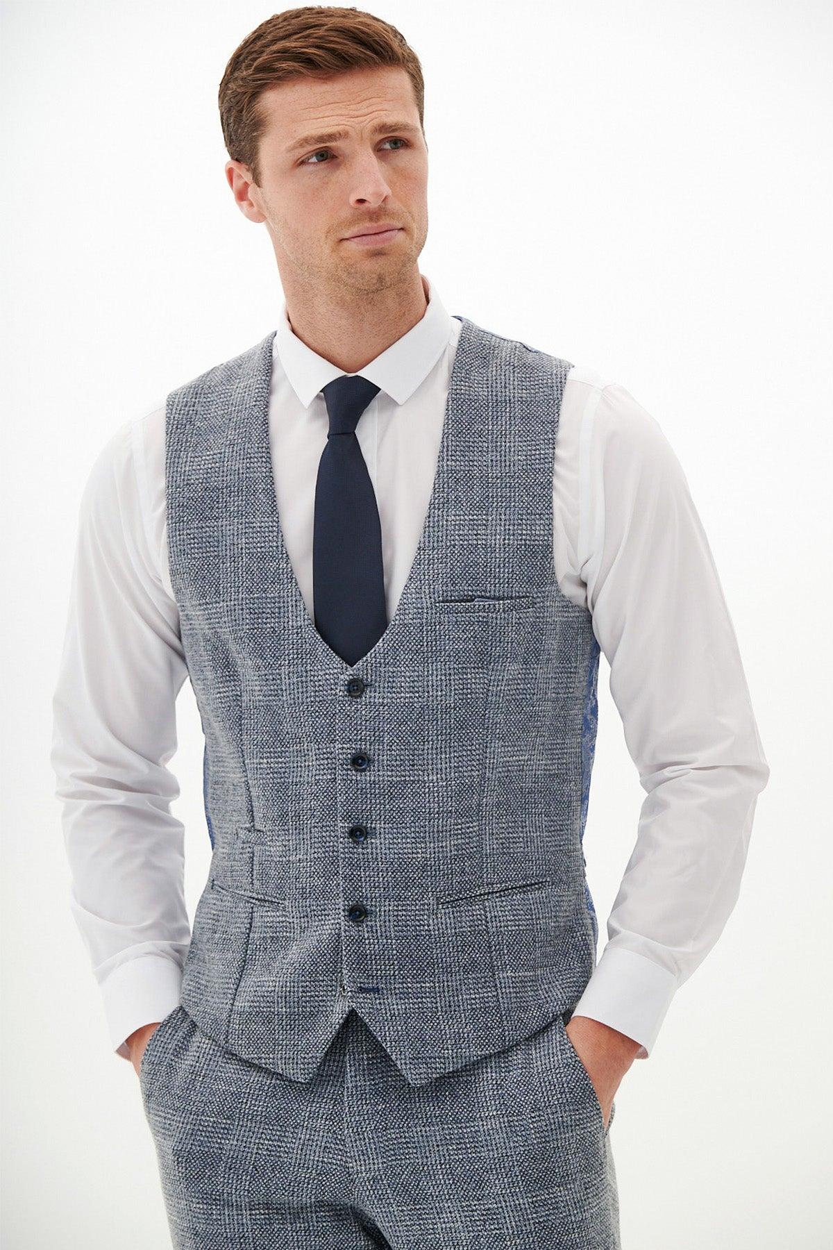 A man in a Marc Darcy KRIS Sky Blue Tweed Waistcoat, white dress shirt, and navy tie gazes away from the camera with his hands in his vest pockets.