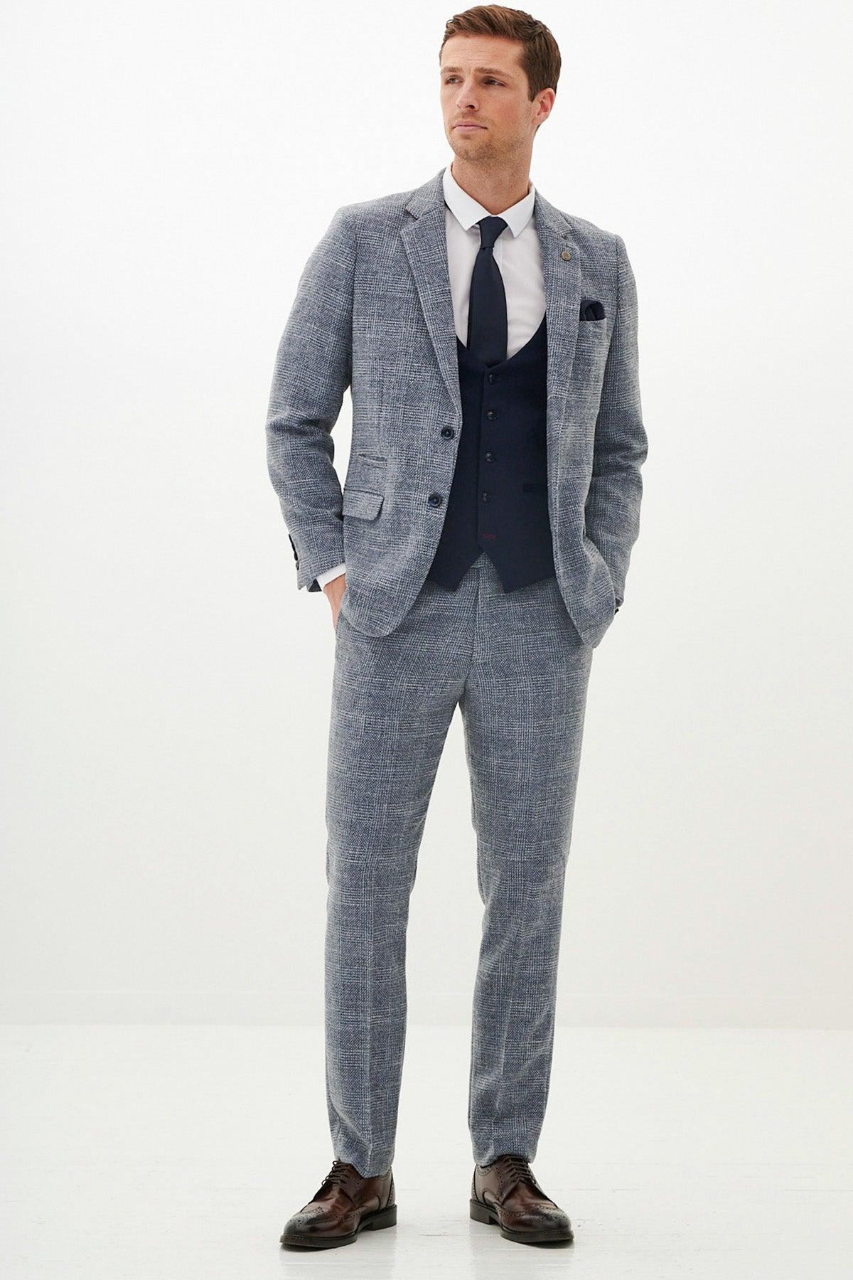 A man stands against a white background in a marcdarcy KRIS Sky Blue Tweed Suit with a Kelvin Navy Waistcoat, crisp white shirt, matching tie, brown dress shoes, and his right hand in his pocket.