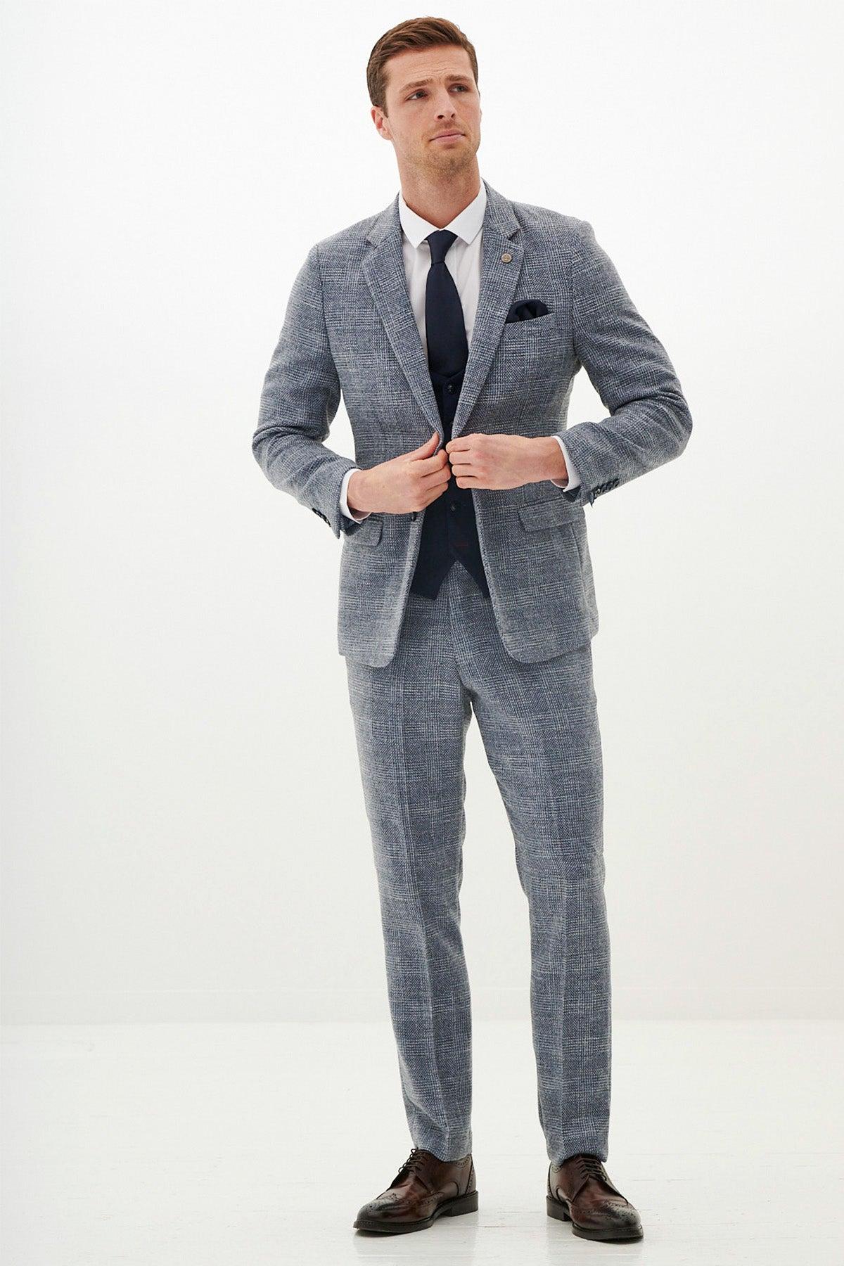 A man wears the marcdarcy KRIS Sky Blue Tweed Suit with a Kelvin Navy Waistcoat, white shirt, navy tie, and brown dress shoes. He adjusts his jacket while standing against a plain white background, looking slightly to the side.