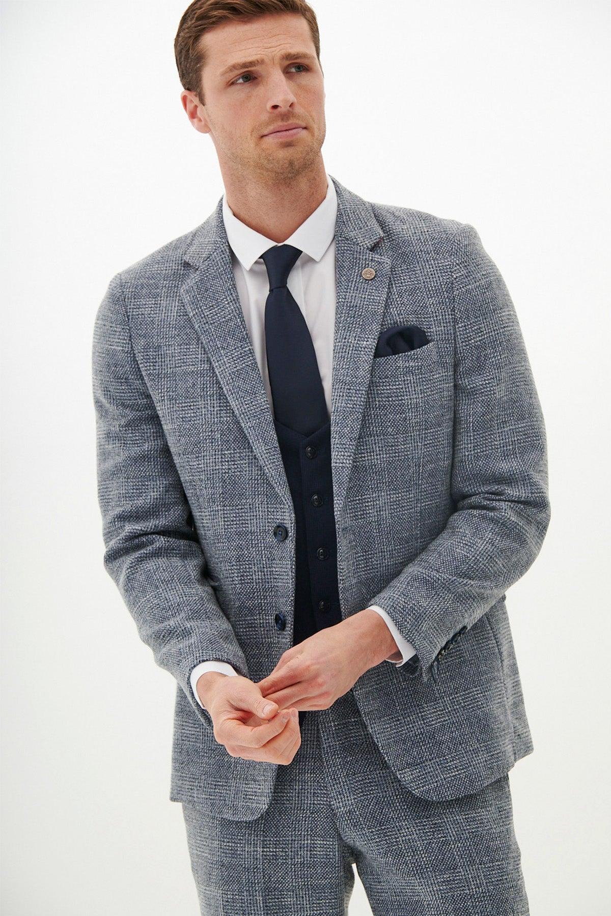 A man wears a marcdarcy KRIS Sky Blue Tweed Suit with a Kelvin Navy Waistcoat and a white shirt, his hands clasped in front of him.
