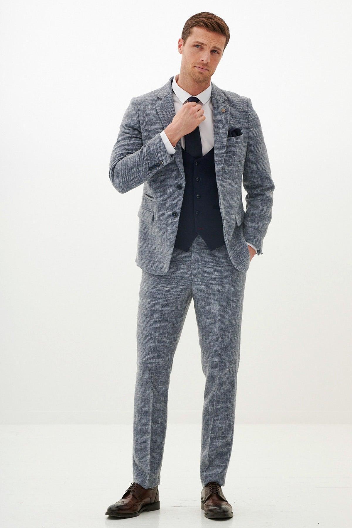 A man in a marcdarcy KRIS Sky Blue Tweed Suit with Kelvin Navy Waistcoat, white shirt, dark tie, brown shoes, and a pocket square adjusts his tie.