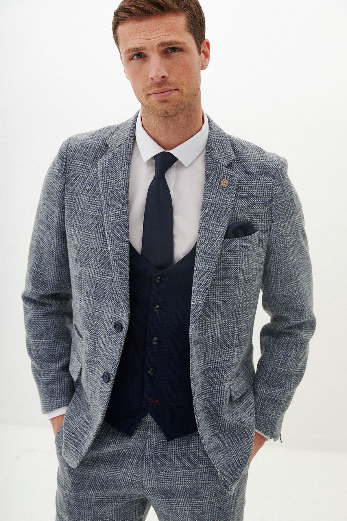 A man in a marcdarcy KRIS sky blue tweed suit with a Kelvin navy waistcoat, white shirt, and navy tie poses with his hands in his pockets.