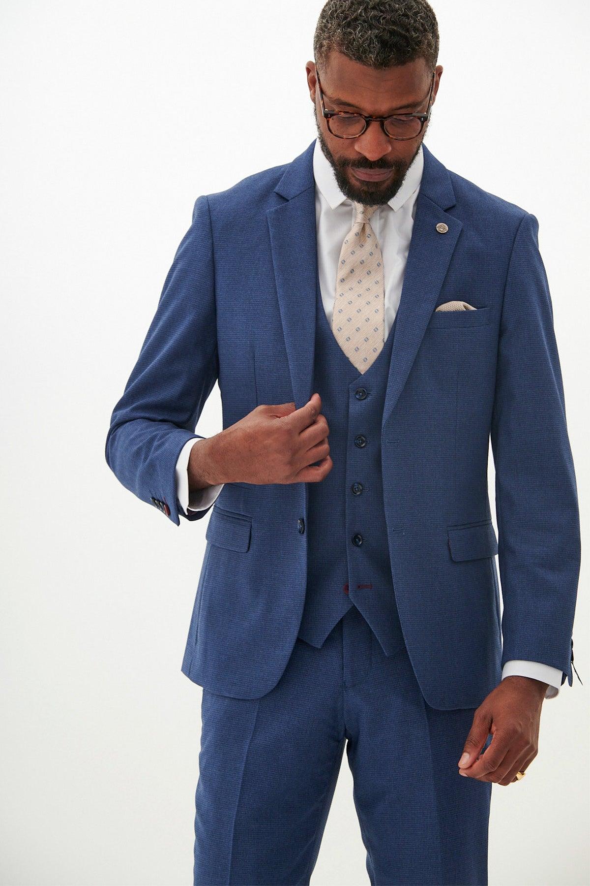 A man wearing the Marc Darcy ASHLEY Navy Blue Blazer three-piece suit with a white shirt, patterned tie, and pocket square stands against a plain white background, looking down and adjusting his jacket.