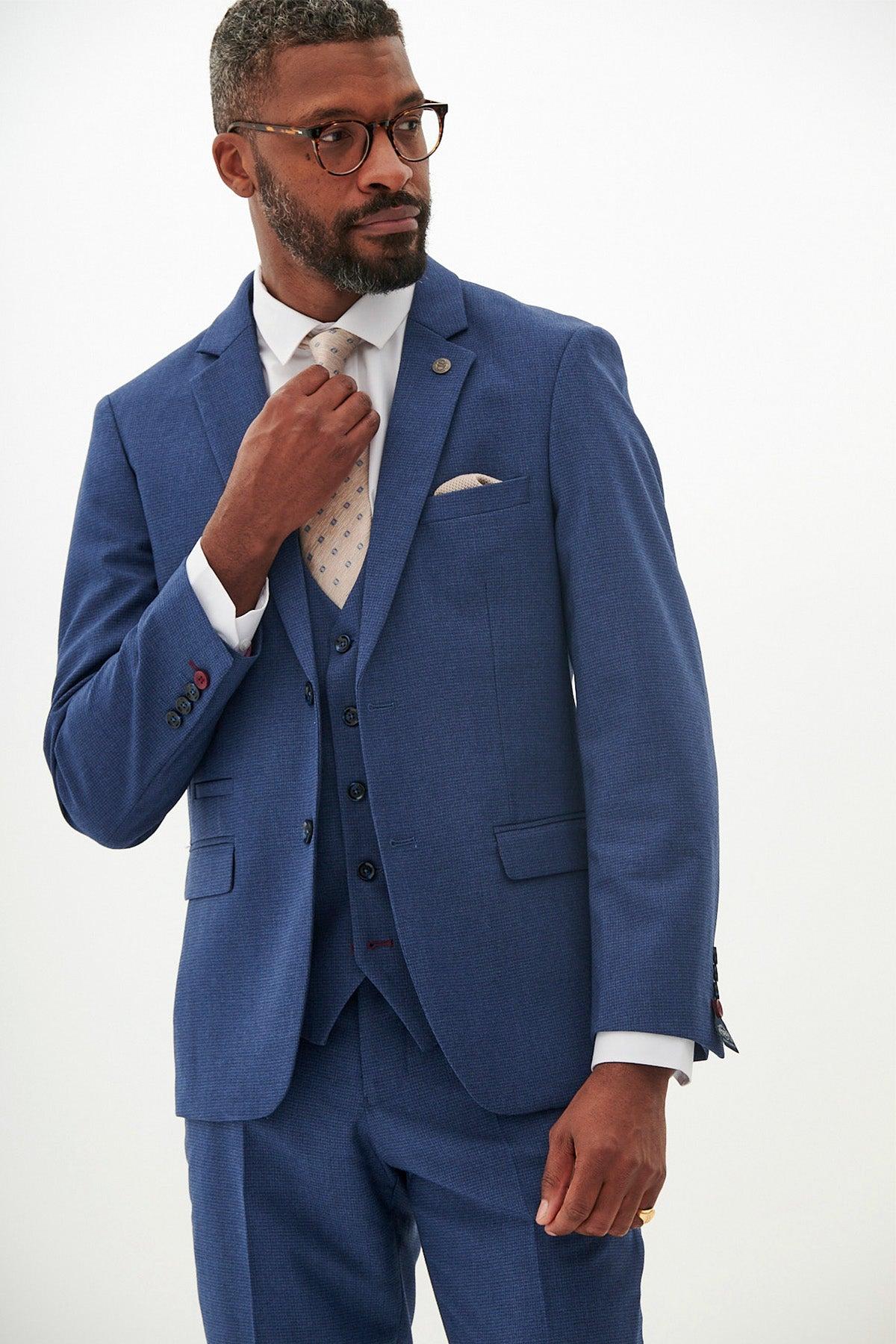 A man in glasses adjusts his tie while wearing the Marc Darcy WHU Collection - ASHLEY Navy Blue Three Piece Suit as worn by Aaron Wan-Bissaka, paired with a white shirt and patterned tie, against a plain light background.