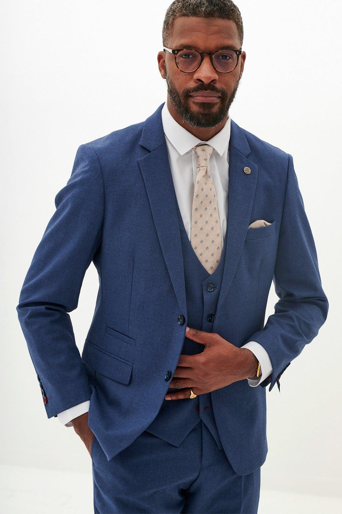A man in a tailored Marc Darcy Everton Collection ASHLEY Navy Blue Three Piece Suit, beige patterned tie, and glasses stands against a plain white background with one hand in his pocket and the other holding his jacket.