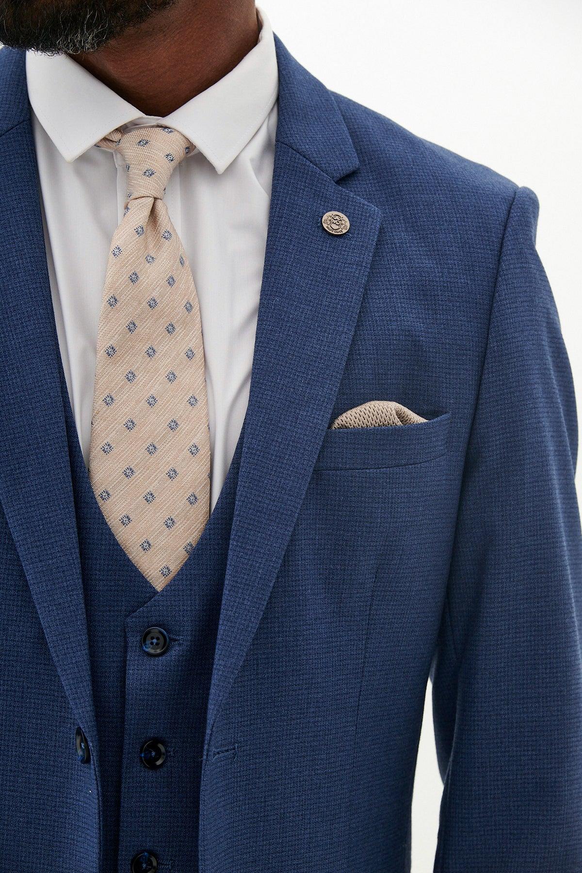 A man wears the Marc Darcy WHU Collection - ASHLEY Navy Blue Three Piece Suit as worn by Aaron Wan-Bissaka, styled with a white shirt, beige patterned tie, matching pocket square, and buttoned jacket with lapel pin over a vest.