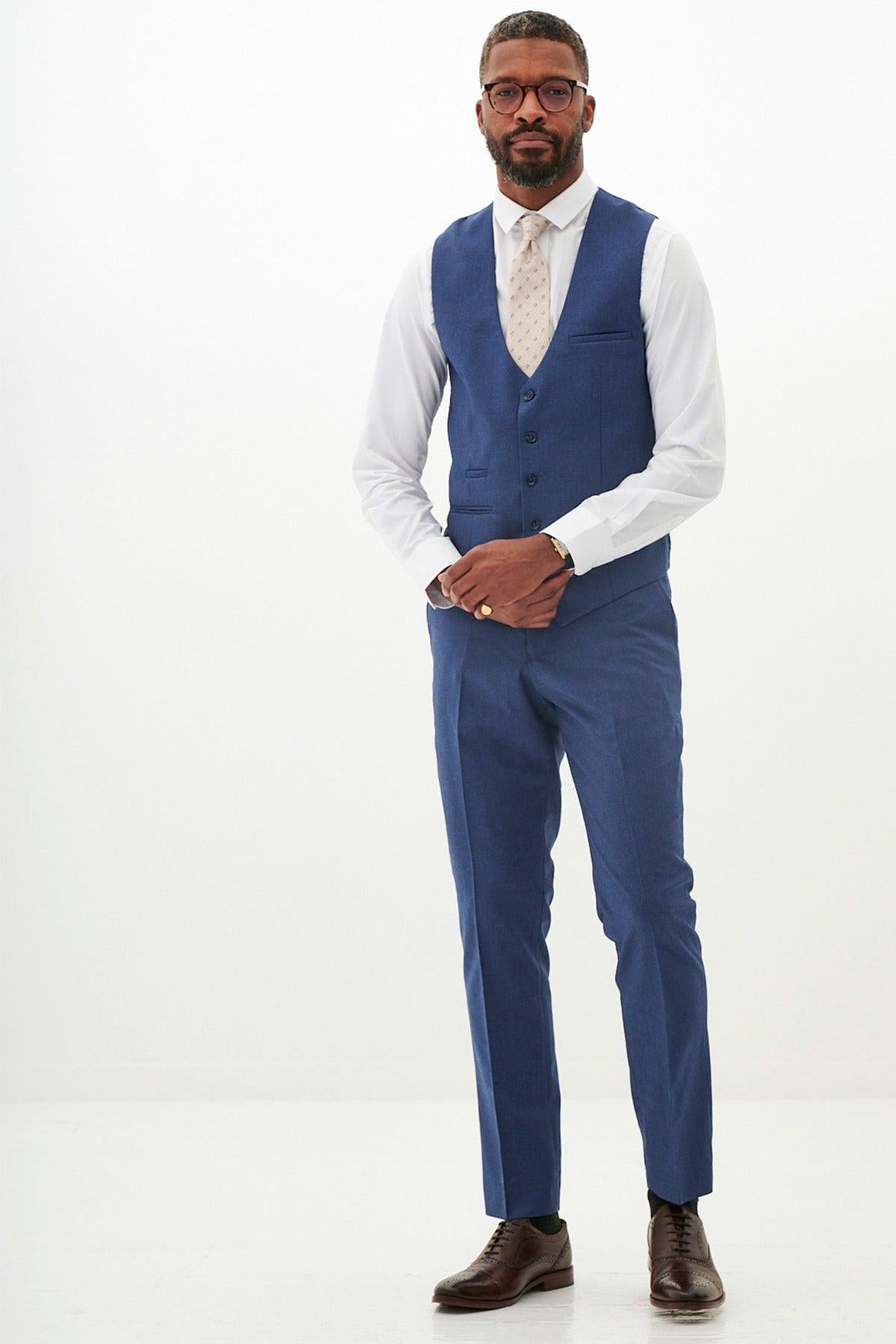 A man in The WHU Collection - ASHLEY Navy Blue Three Piece Suit by Marc Darcy, as worn by Aaron Wan-Bissaka, featuring a blue vest, white shirt, and blue pants—wearing glasses and brown shoes.
