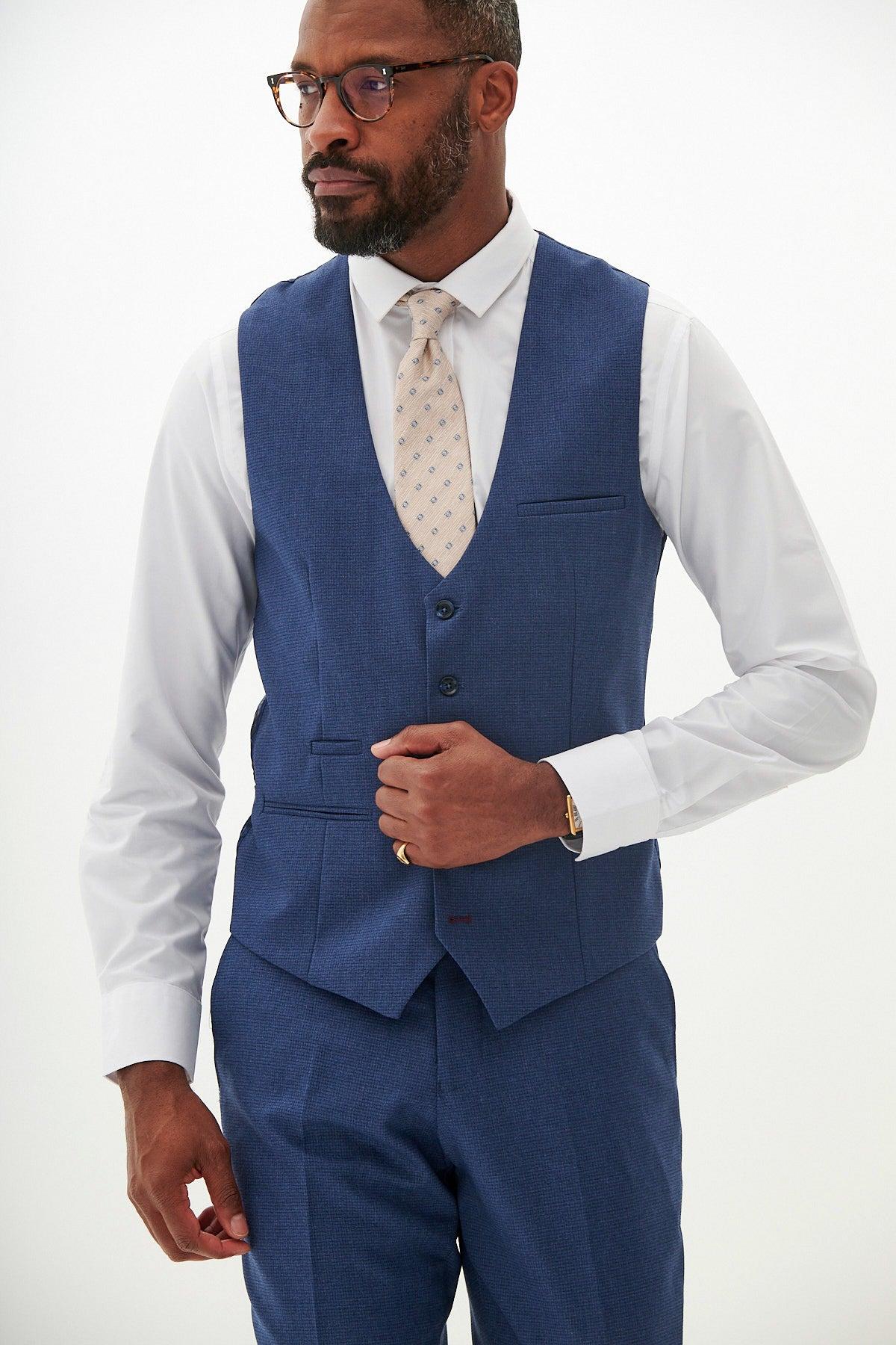 A Black man with glasses wears the ASHLEY Navy Blue Waistcoat by Marc Darcy over a white shirt and patterned tie, showing off a gold ring and watch as he rests his hand on the formal waistcoat.