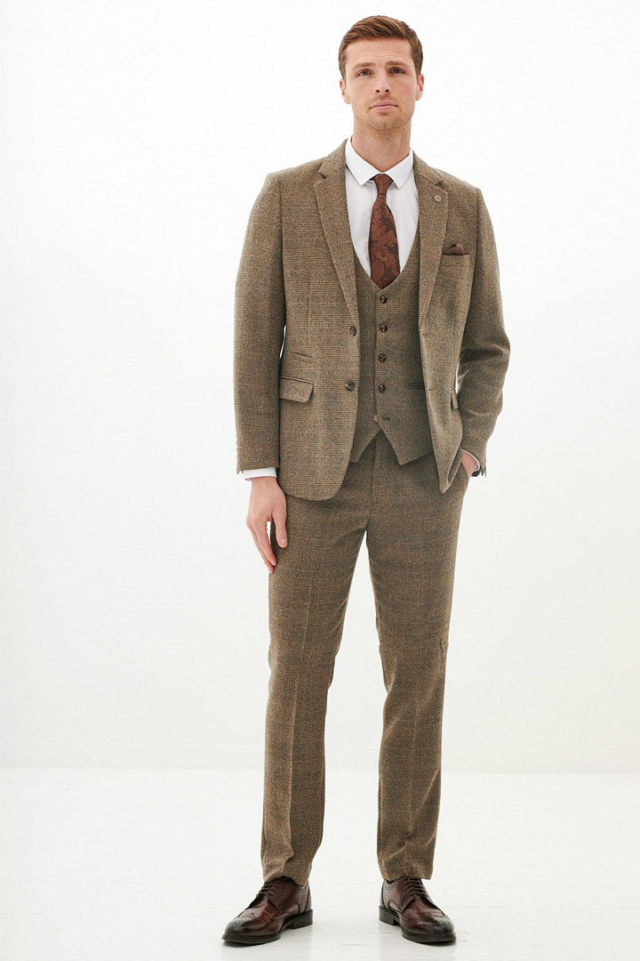 A man with short hair and a neutral expression stands against a white background, wearing the marcdarcy KRIS Tan Tweed Three Piece Suit, a white dress shirt, patterned brown tie, and brown dress shoes, with one hand in his pocket.