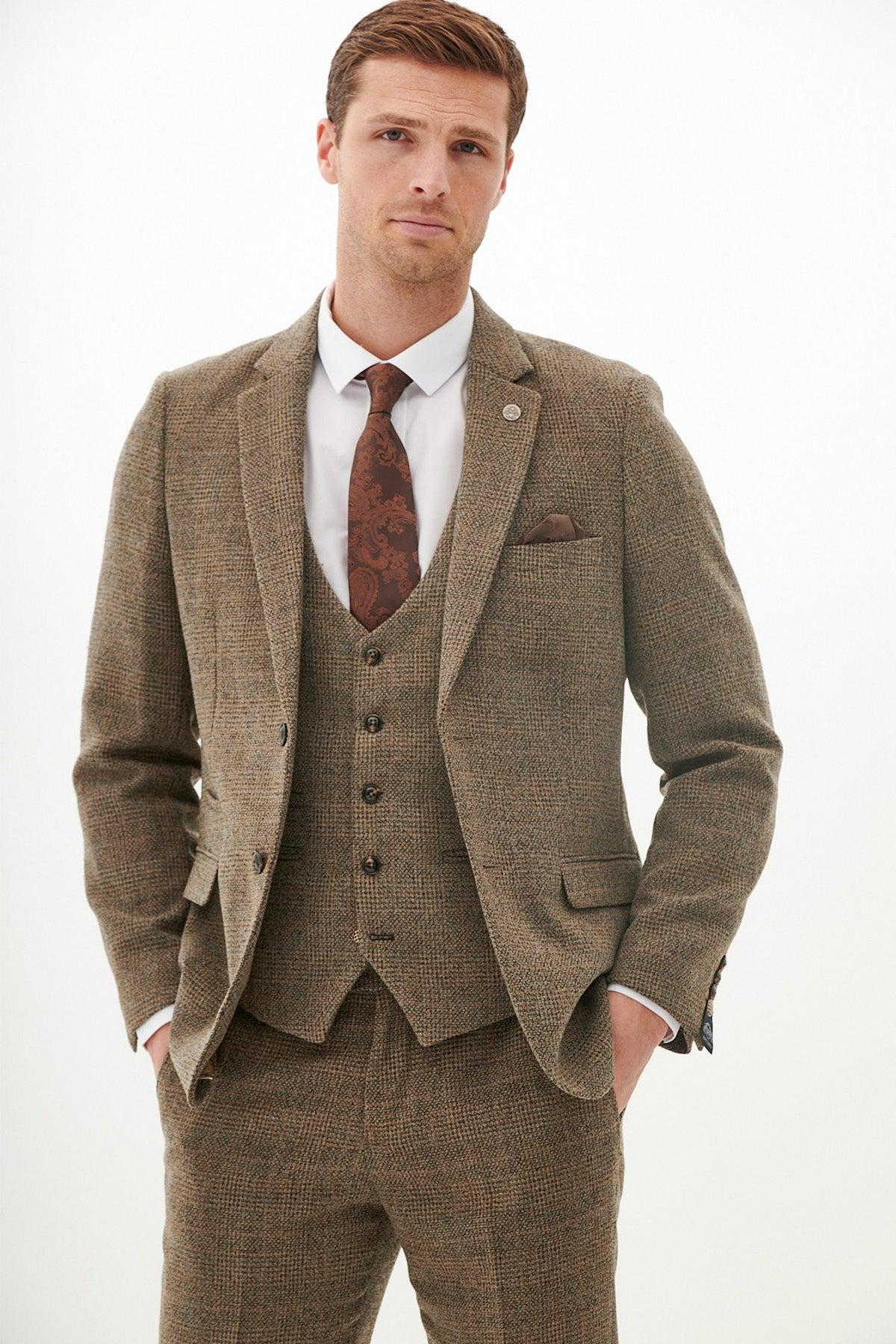 A man models the marcdarcy KRIS - Tan Tweed Three Piece Suit with a white shirt and brown patterned tie, standing with hands in pockets against a plain white background.