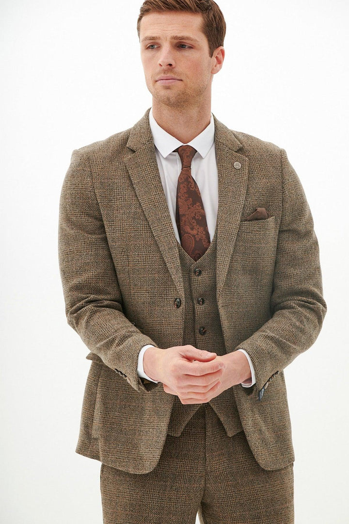 A man wearing the marcdarcy KRIS - Tan Tweed Blazer, a white dress shirt, and a patterned brown tie stands with hands clasped, gazing slightly to the side against a plain white background.