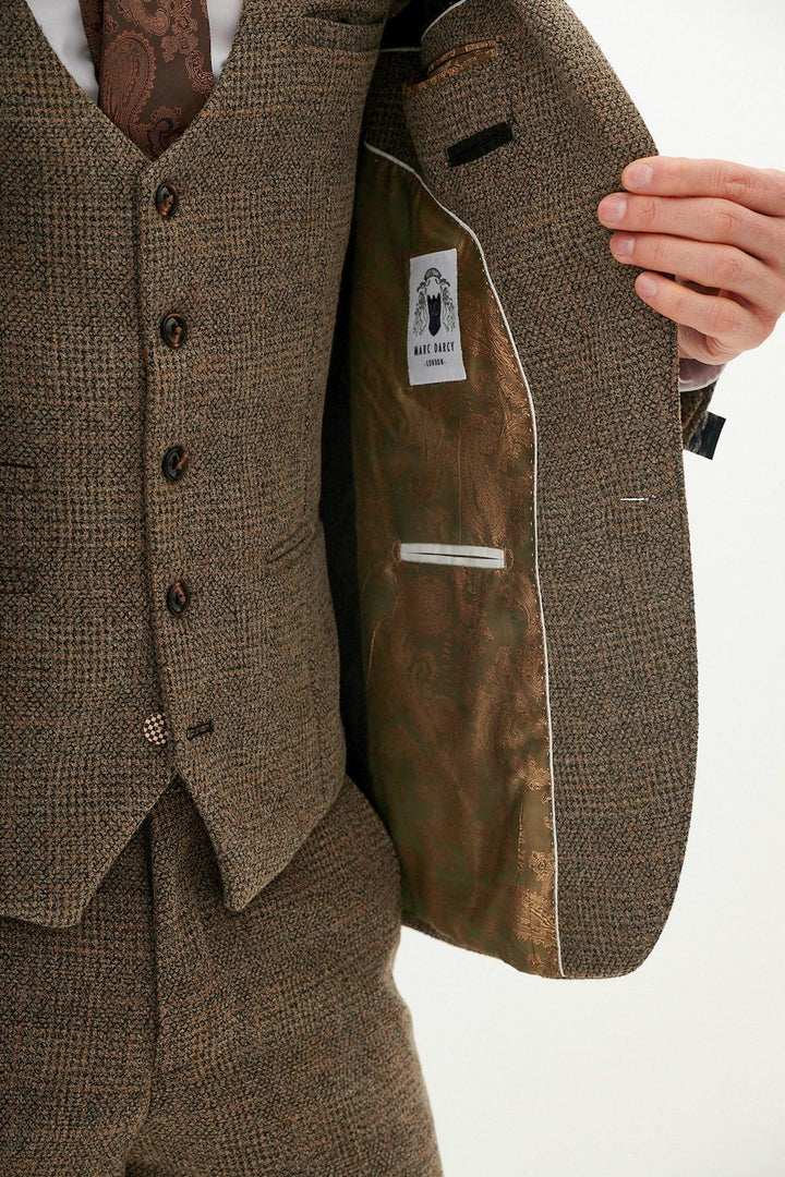 A man in a slim fit marcdarcy KRIS tan tweed three piece suit holds open his jacket, showing its patterned brown lining and brand label. The suit includes a vest, matching trousers, and a paisley brown tie.