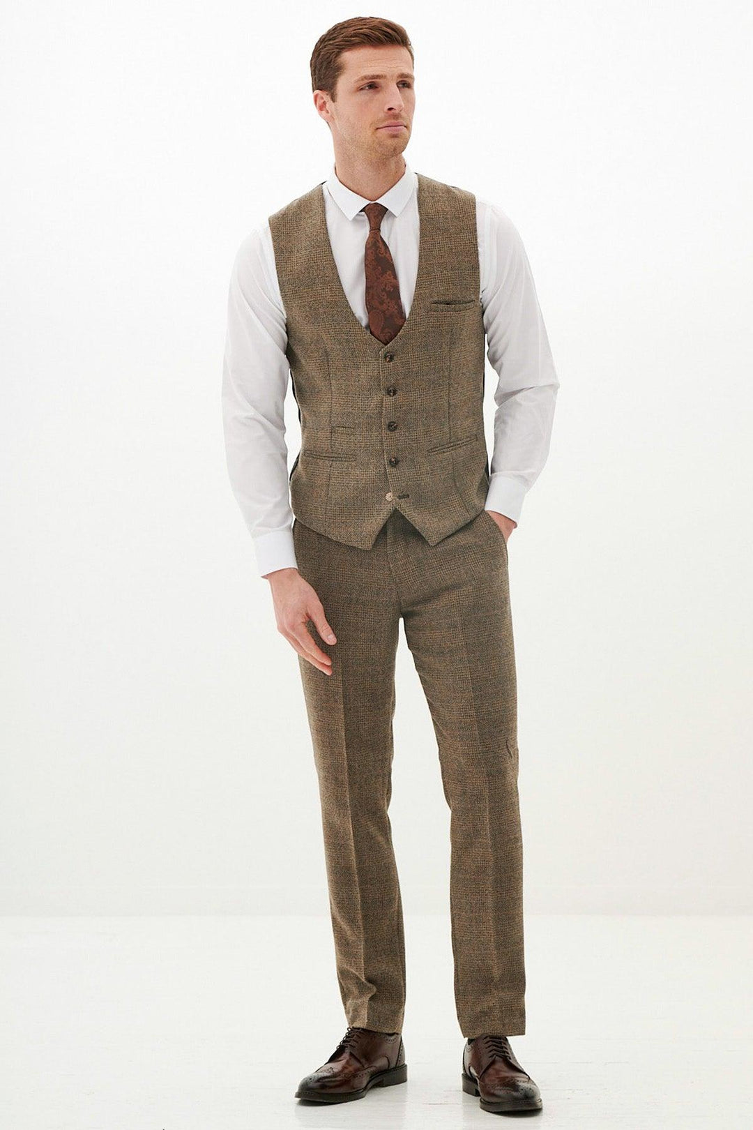 A man poses against a white background wearing the marcdarcy KRIS Tan Tweed Three Piece Suit, styled with a white shirt, brown patterned tie, matching waistcoat and trousers, and dark brown dress shoes.
