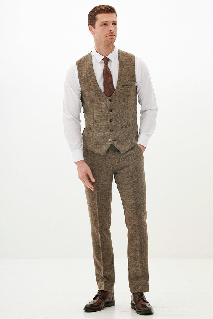 A man with short hair and a neutral expression stands against a white background, wearing the Marc Darcy KRIS Tan Tweed Waistcoat with matching trousers, a white shirt, brown patterned tie, and dark brown dress shoes.