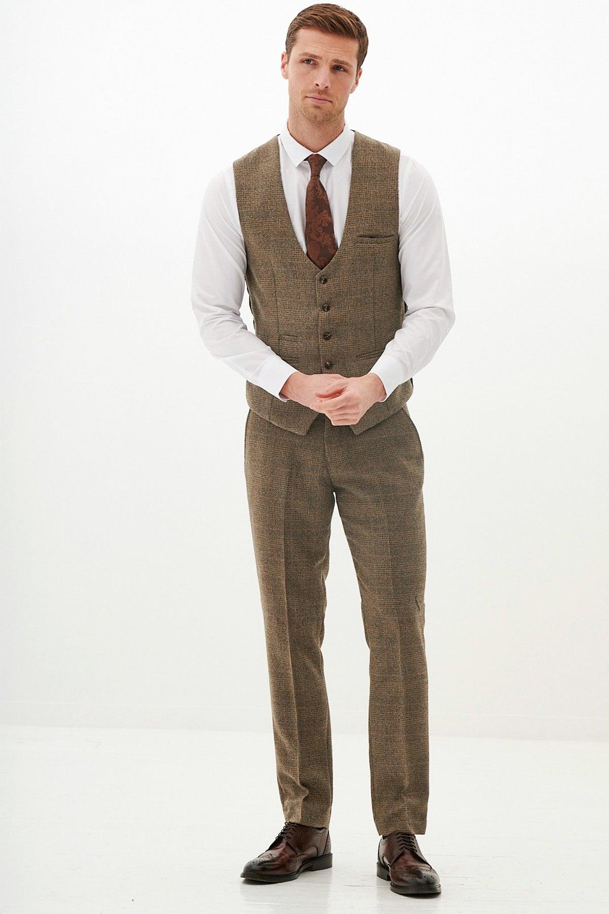 A man stands against a plain white background, wearing a Marc Darcy KRIS tan tweed waistcoat with matching trousers, a white dress shirt, brown tie, and brown dress shoes. He has short hair and a neutral expression.