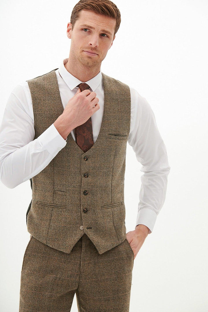 A man wearing the Marc Darcy KRIS - Tan Tweed Waistcoat stands against a plain light background, adjusting his brown tie and looking at the camera.