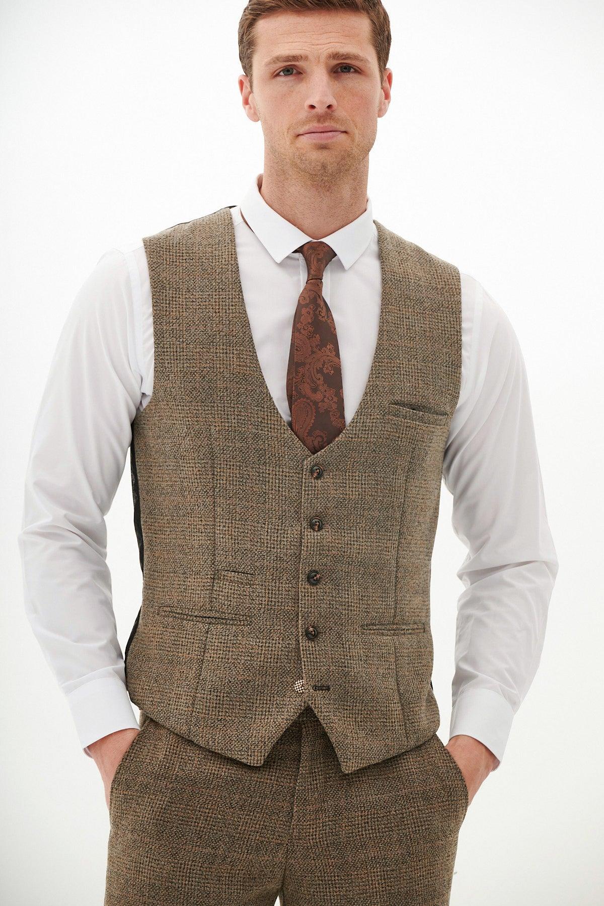 A man wearing the Marc Darcy KRIS Tan Tweed Waistcoat with matching trousers, a white dress shirt, and a patterned brown tie stands with his hands in his pockets against a plain, light background.