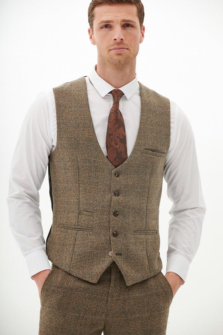 A man stands with hands in pockets against a white background, wearing a Marc Darcy KRIS Tan Tweed Waistcoat with matching trousers, a white dress shirt, and a brown patterned tie.