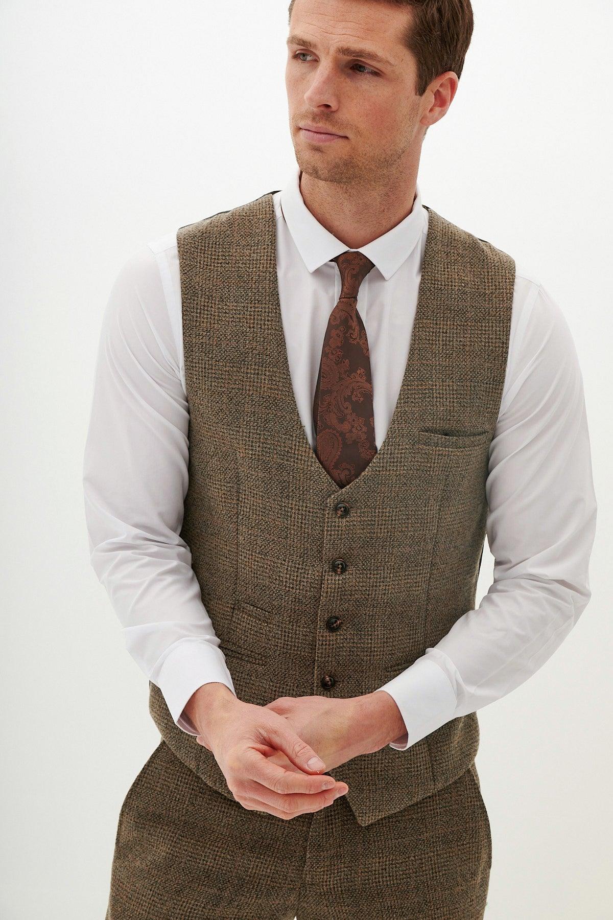 A man wearing the Marc Darcy KRIS Tan Tweed Waistcoat, matching trousers, a white dress shirt, and a patterned brown tie stands against a plain white background, looking to the side with his hands clasped.