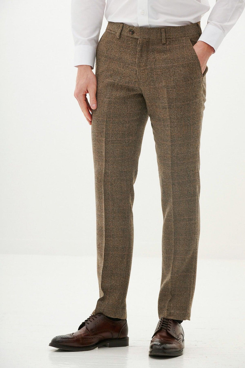 A man wears marcdarcy KRIS tan tweed trousers with a white shirt and brown wingtip shoes against a white background.