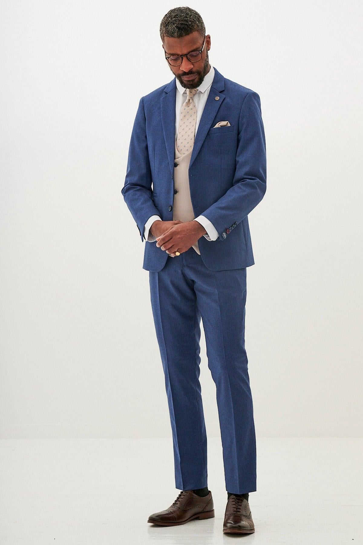 A man models the Marc Darcy ASHLEY - Navy Blue Suit with Kelvin Stone Waistcoat, styled with a white shirt, light tie, and brown shoes, standing against a plain white background with his hands clasped as he looks down.
