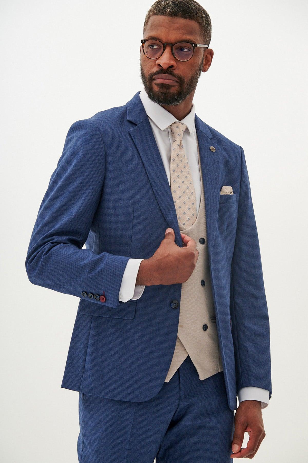 A man poses against a white background wearing the Marc Darcy ASHLEY Navy Blue Suit with Kelvin Stone Waistcoat, paired with a white shirt, patterned cream tie, and round tortoiseshell glasses.