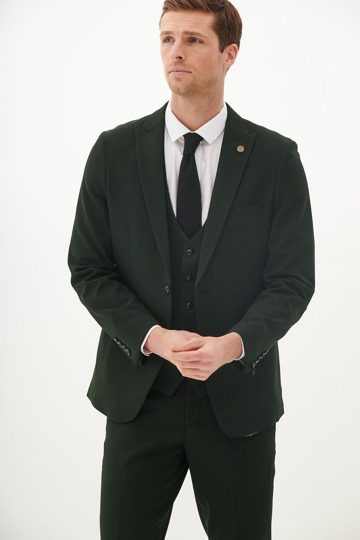 A man wears the Marc Darcy Leicester City F.C. Collection - BROMLEY Olive Green Check Suit as worn by Harry Winks, paired with a white shirt and black tie, standing against a white background with his hands clasped.