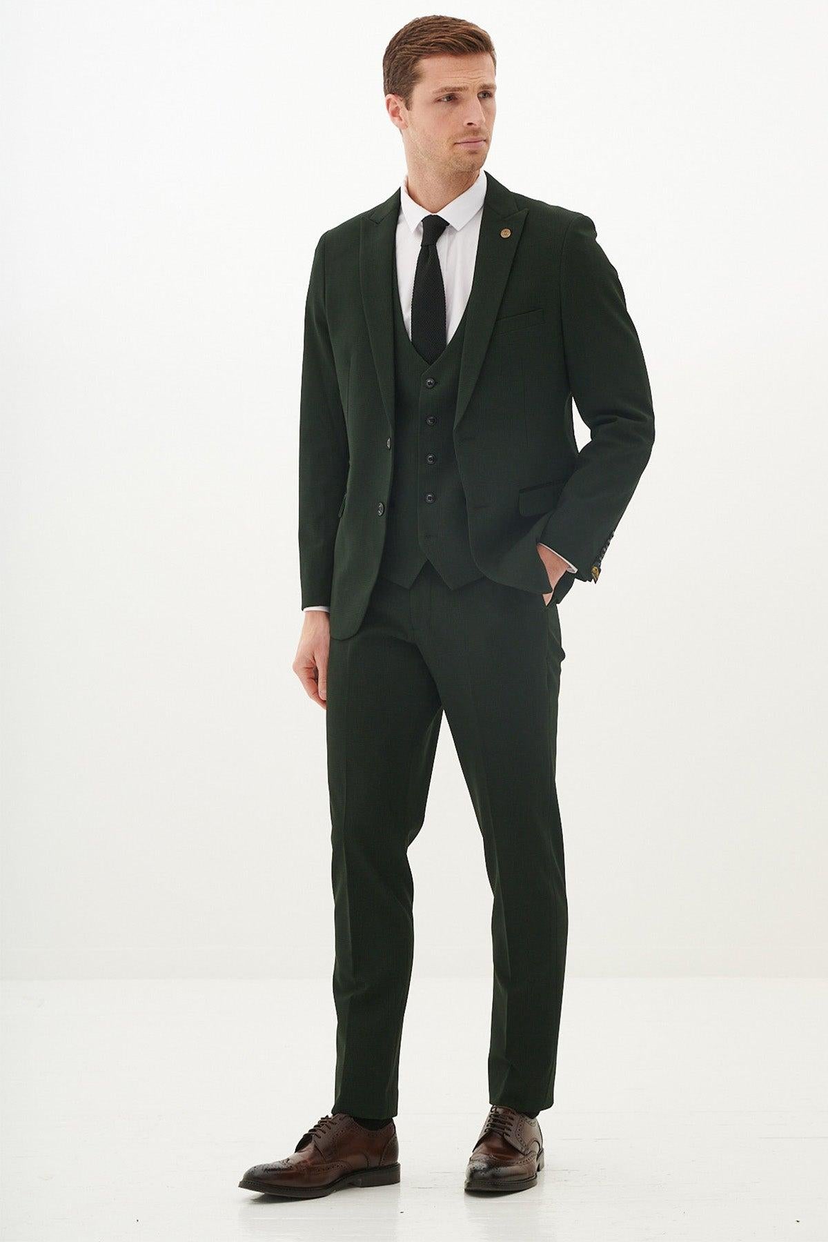 A man models the Marc Darcy BROMLEY - Olive Green Check Three Piece Suit with a white dress shirt, black tie, and brown dress shoes, standing against a white background and looking left with his hand in his pocket.