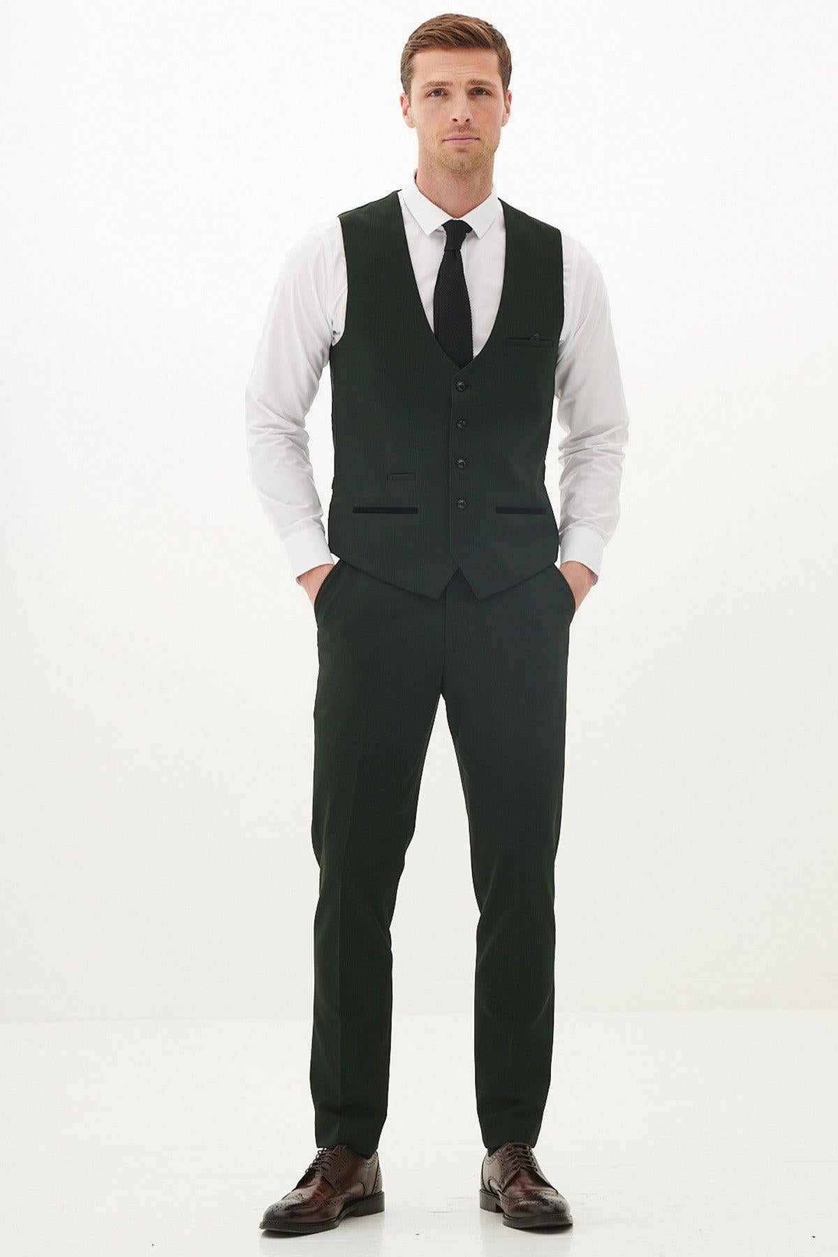 A man stands with his hands in his pockets, wearing the Marc Darcy BROMLEY Olive Green Check Three Piece Suit—a tailored set with a vest, Prince of Wales check pants, black tie, white shirt, and brown dress shoes.