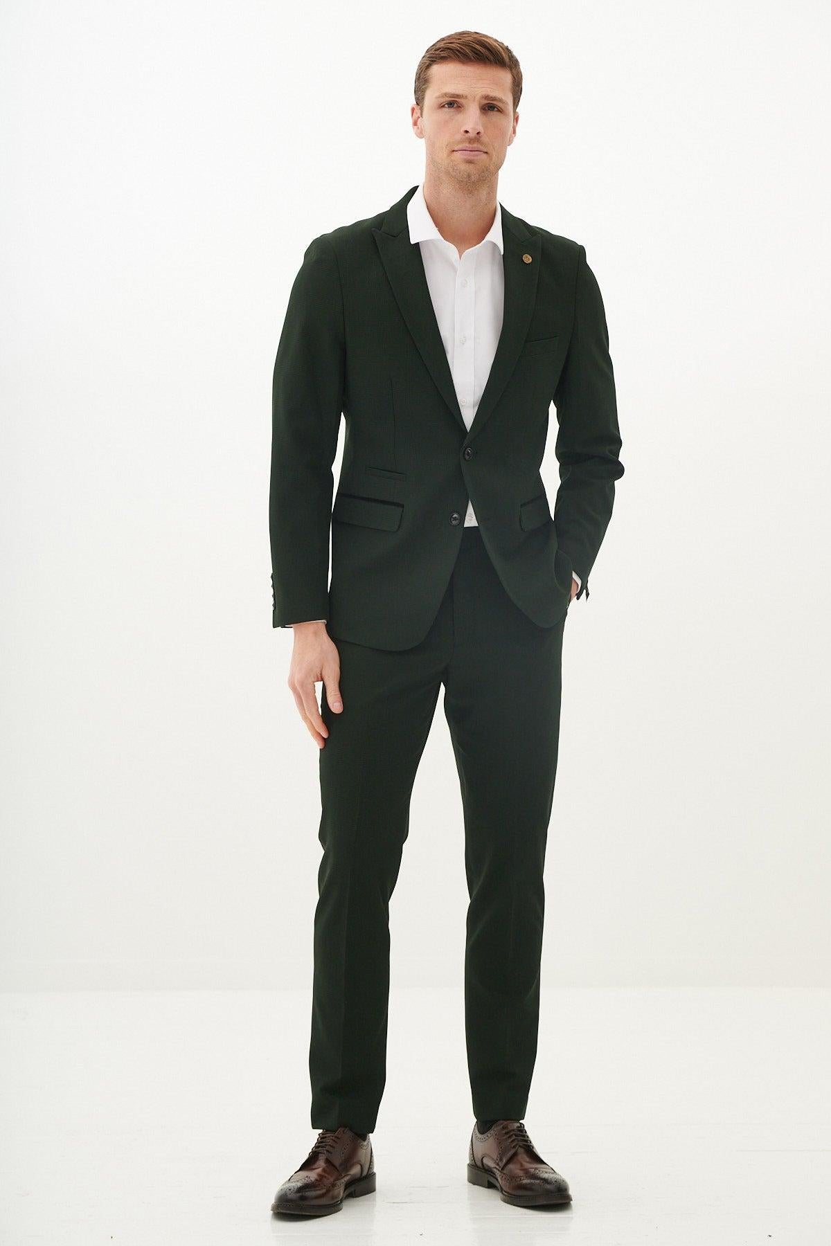 A man stands against a white background wearing the Marc Darcy Cardiff City F.C. Collection BROMLEY olive green check blazer as worn by Alex Robertson, paired with a white shirt, gold lapel pin, and brown leather brogues.
