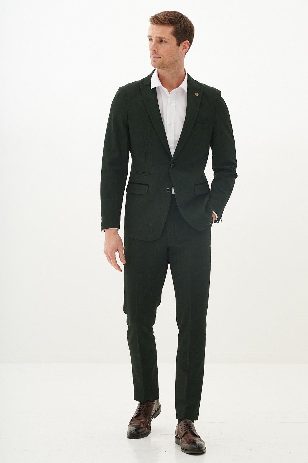 A man stands against a white background wearing a slim-fit white shirt, brown leather brogue shoes, and the Marc Darcy Cardiff City F.C. Collection - BROMLEY Olive Green Check Blazer as worn by Alex Robertson.
