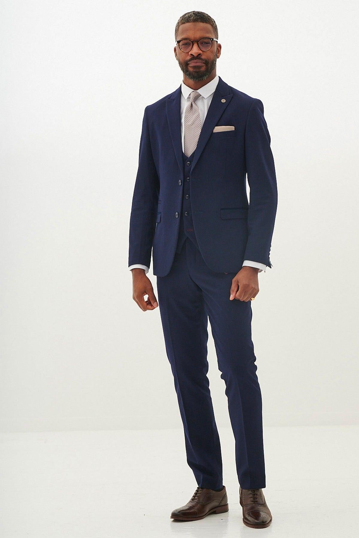 A man with a neat beard and short hair wears the Marc Darcy BROMLEY Navy Check Three Piece Suit with a white shirt, light patterned tie, matching pocket square, brown shoes, and glasses against a plain white background.