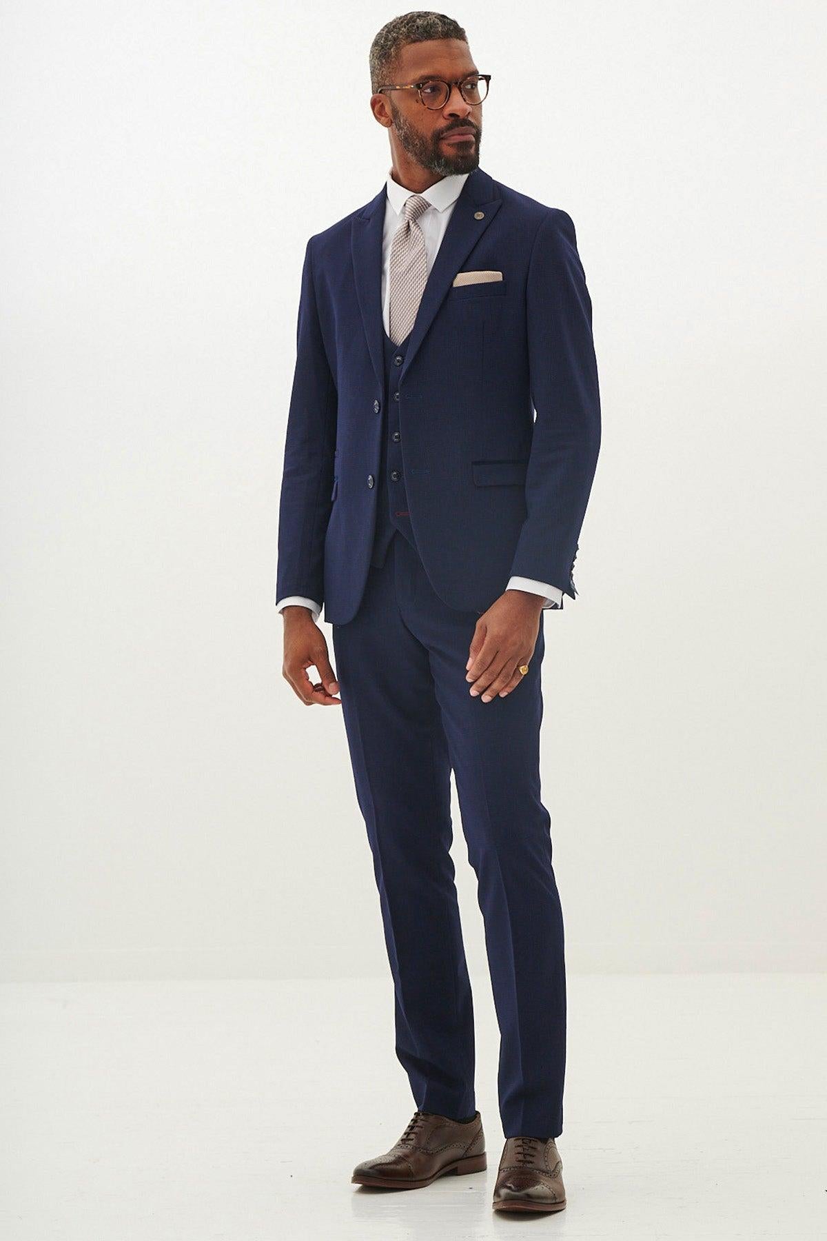 Man wearing the Marc Darcy BROMLEY Navy With Monty Waistcoat Suit—as worn by Jarrod Bowen—with a crisp white shirt, patterned tie, and brown dress shoes, looking to the side.