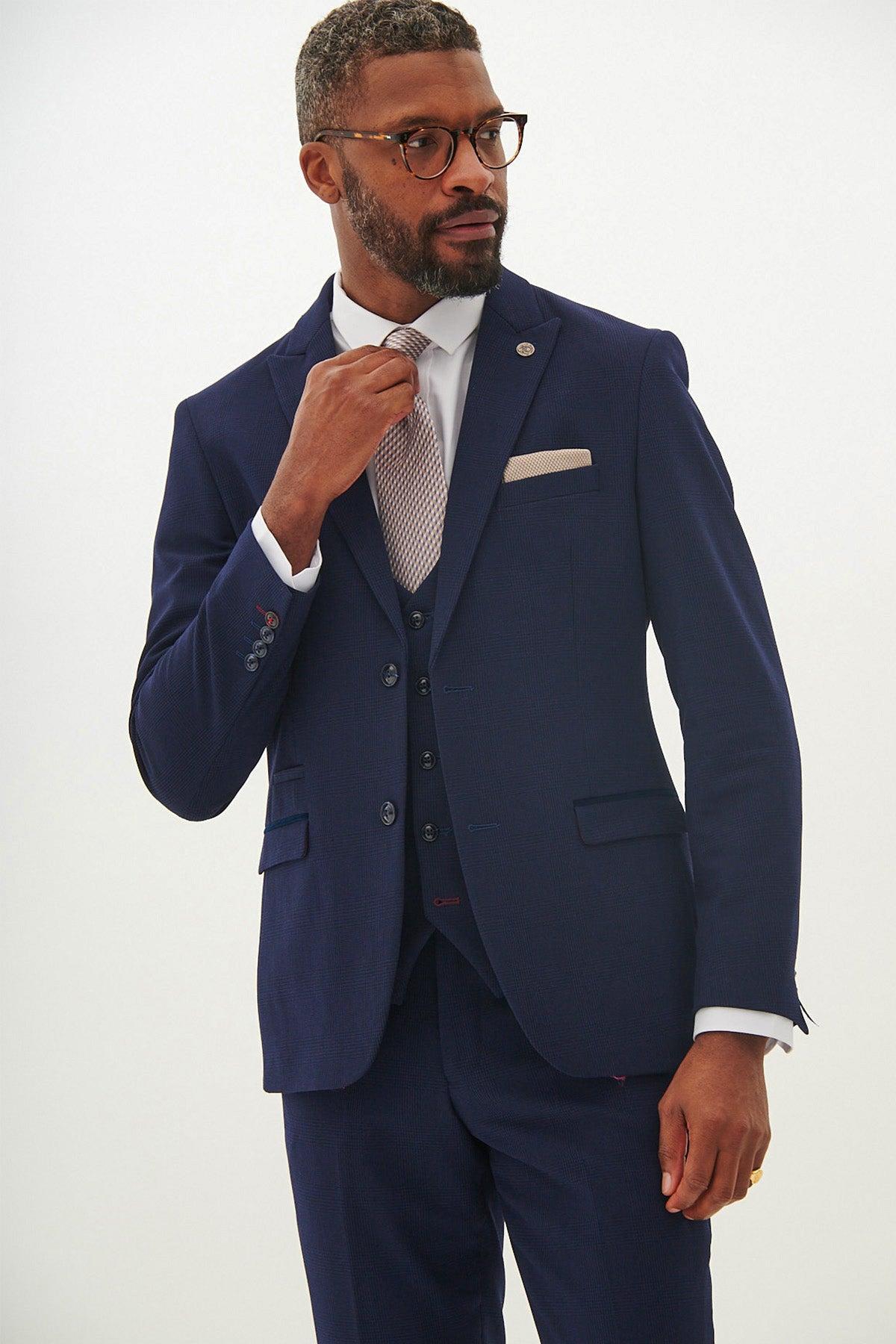 A man with short hair and a beard wears glasses and the Marc Darcy BROMLEY - Navy Check Three Piece Suit, featuring a vest, white shirt, and patterned tie. He adjusts his tie with a calm, confident expression against a plain light background.