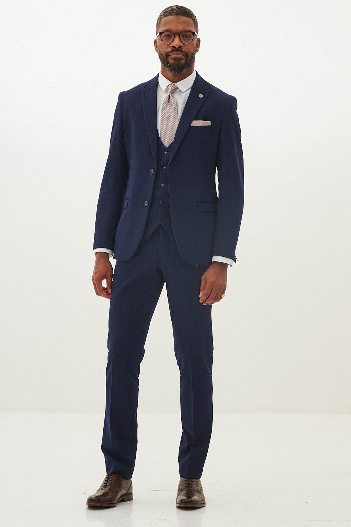 A man with short hair, a beard, and glasses poses confidently in the BROMLEY - Navy Check Three Piece Suit by Marc Darcy, paired with a white shirt, light tie, and brown dress shoes against a plain background.